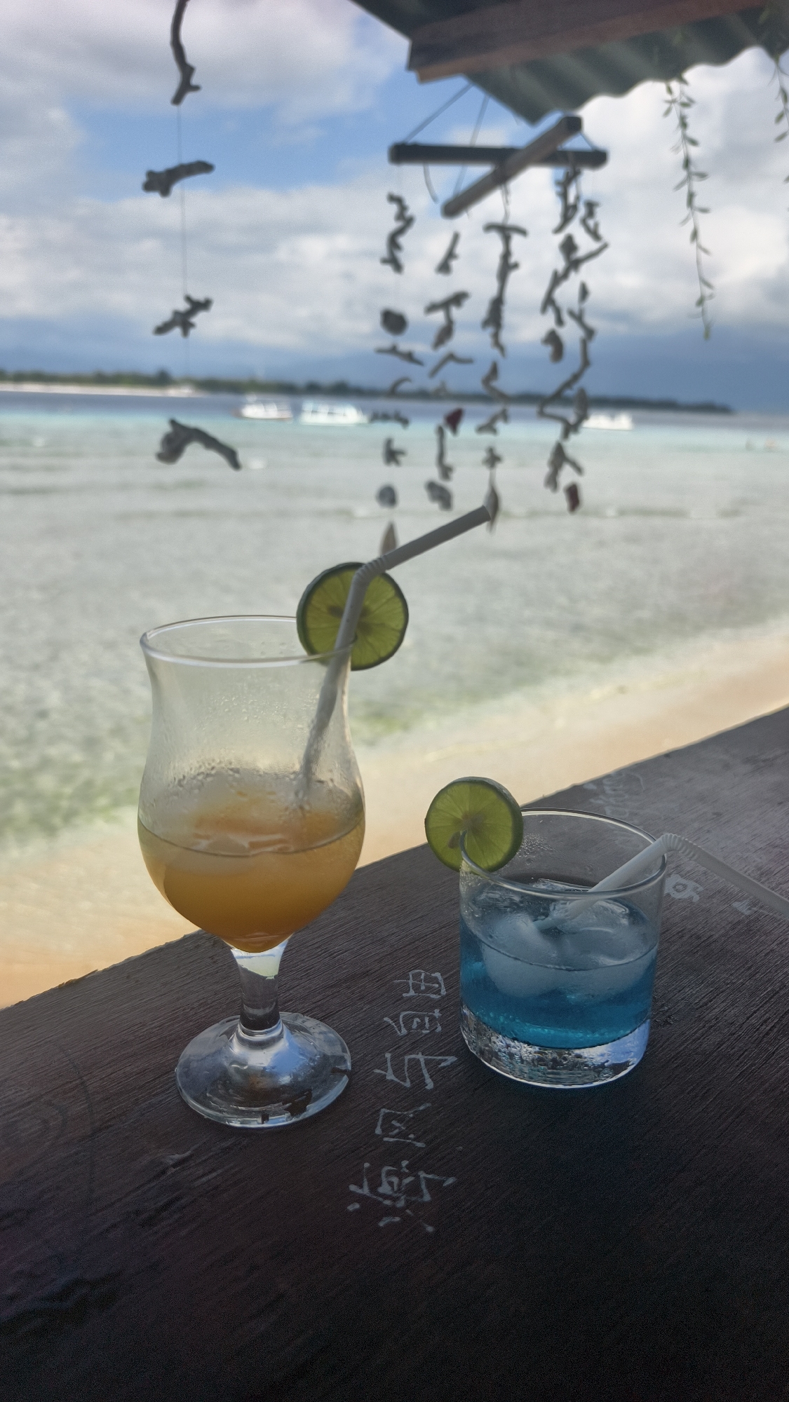 A close-up shot shows two beverages on a dark, wooden surface with faded, white lettering in an unknown language. The drink on the left is a tall glass filled with a golden-orange liquid, garnished with a lime slice and a straw. To the right, a shorter glass holds a blue liquid with ice cubes, also garnished with a lime slice and a straw. The backdrop features a beach scene with a calm, shallow sea, distant boats, and a coastline under a partly cloudy sky. Hanging decorations dangle from an unseen structure above, adding to the tropical, outdoor ambiance. This scene is captured in an unknown city in Indonesia.