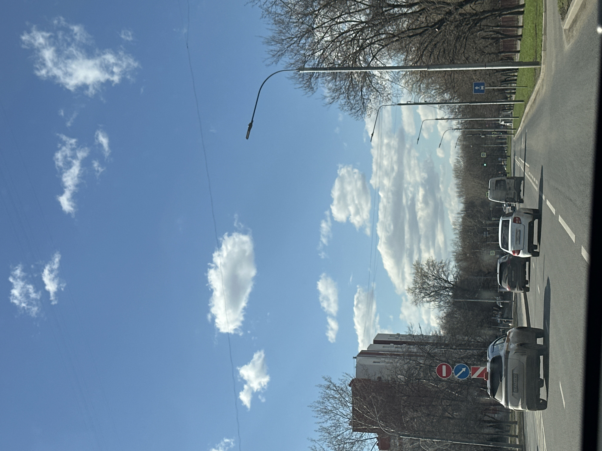 The image depicts a street scene in Moscow, Russia, on a sunny day with scattered clouds. The view is from inside a vehicle looking out onto a road with several cars traveling in the same direction. On the right side of the road, there are trees lining the street, streetlights, and buildings. A traffic sign with a "no entry" symbol crossed with a blue arrow indicating the direction of traffic is visible. The sky is blue with white clouds. The image is shot at an angle.
