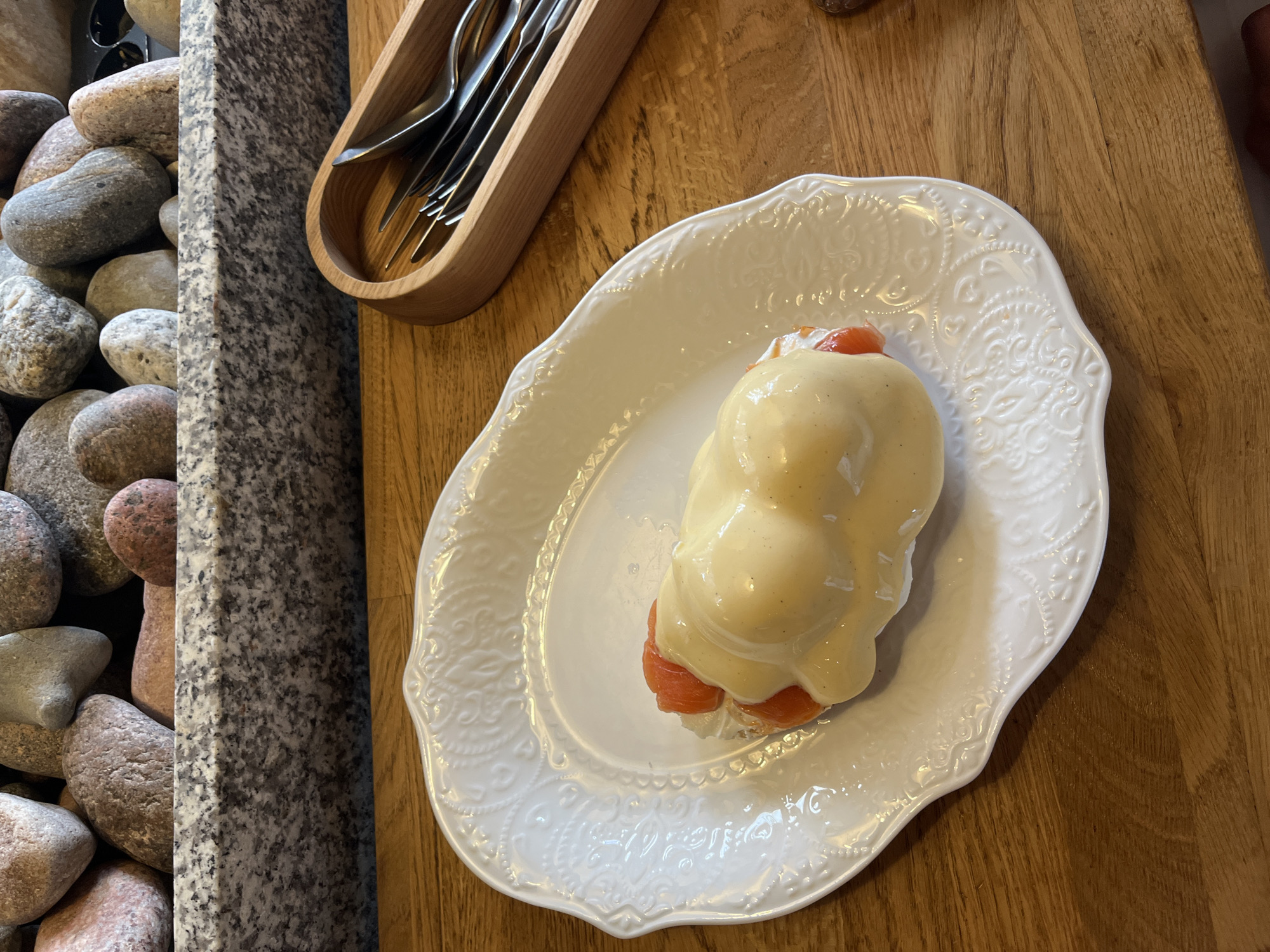 A single serving of eggs Benedict with salmon is presented on a white, ornate plate. The dish is topped with a generous amount of hollandaise sauce and the salmon is visible peeking through. The plate rests on a wooden surface, possibly a table. In the background, a collection of decorative stones and a granite ledge are visible. To the right, a wooden holder contains cutlery, including forks and spoons. The overall scene suggests a restaurant setting. The location is Zelenogradsk, Russia.