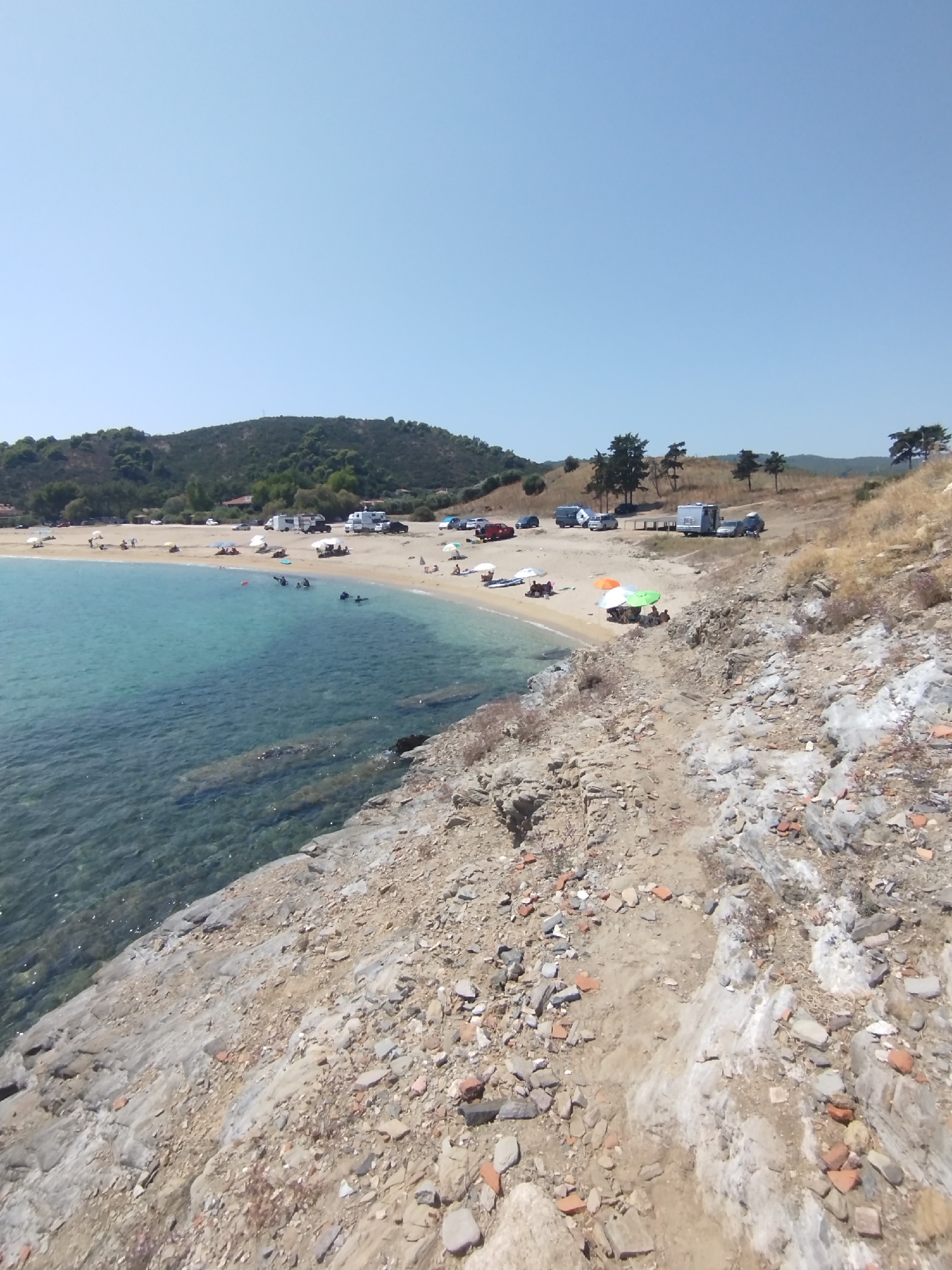 The image depicts a scenic view of a coastal area with a clear blue sky above. In the foreground, a rocky, uneven terrain leads down to a calm, turquoise body of water. The water is shallow near the shore, revealing submerged rocks and the sandy seabed.
Beyond the water, a sandy beach is visible, populated with several beachgoers. Some are swimming or wading in the water, while others are relaxing on the sand under beach umbrellas. A few tents and recreational vehicles are parked along the edge of the beach, suggesting a campsite or a day-use area.
Further inland, a gently sloping hill covered with sparse vegetation and trees rises in the background. The overall environment suggests a tranquil beach destination, likely in a Mediterranean climate, consistent with the location in Sithonia Municipal Unit, Greece. The presence of the sea, beach, and surrounding natural landscape indicates a coastal recreational area.