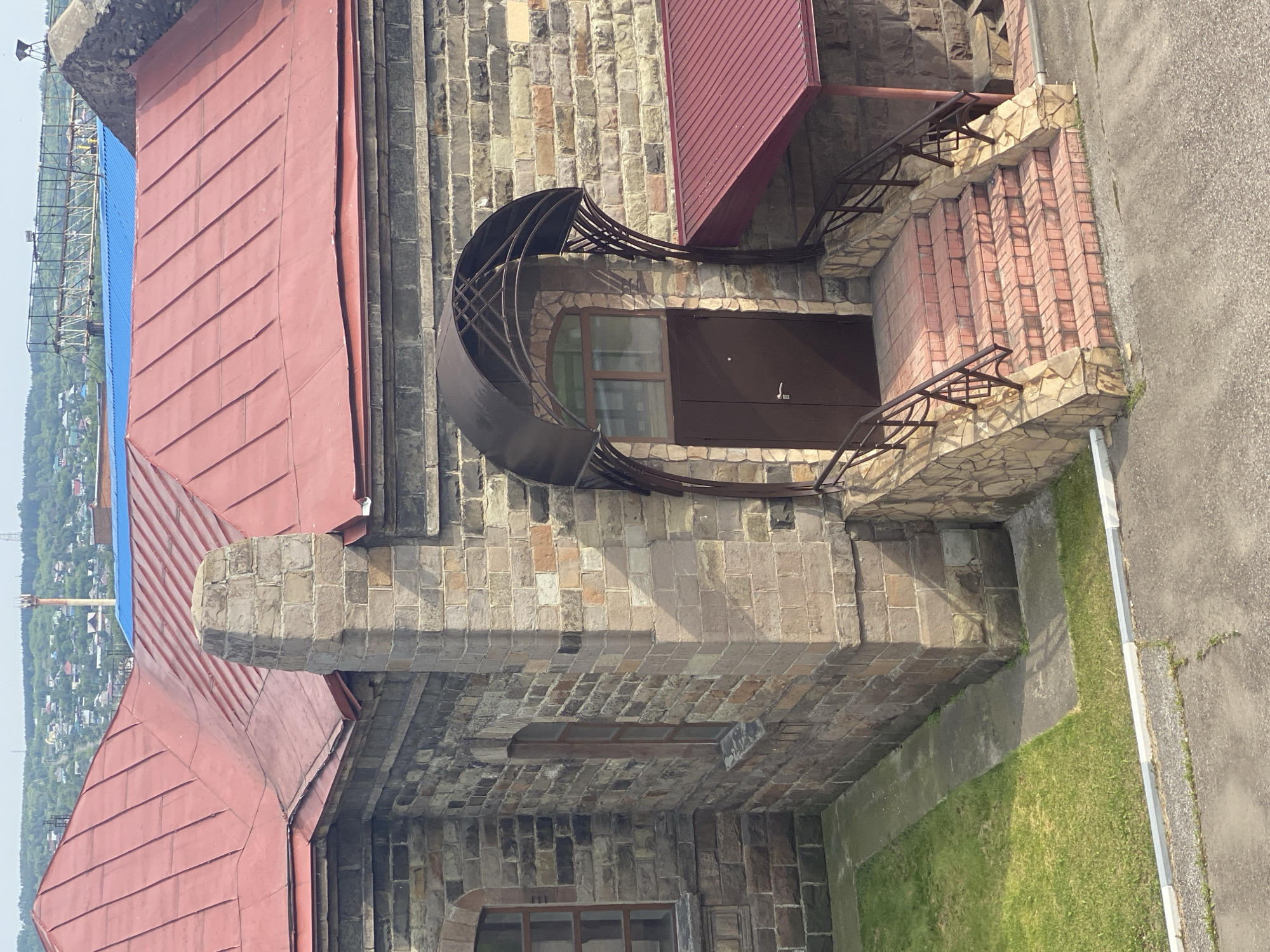 The image shows the exterior of a building constructed with rough-hewn, multi-colored stone blocks. The building features a red, gabled metal roof and a prominent stone chimney. An arched doorway with a brown metal canopy is visible, leading to a set of reddish-pink tiled stairs with metal railings. The building is situated in a landscape with green grass, a paved area, and a view of a town with houses and distant hills under a cloudy sky. The architectural style suggests a historical or industrial structure. The location is identified as Novokuznetsk, Russia, likely an older part of the city given the stone construction. No people are visible in the image.