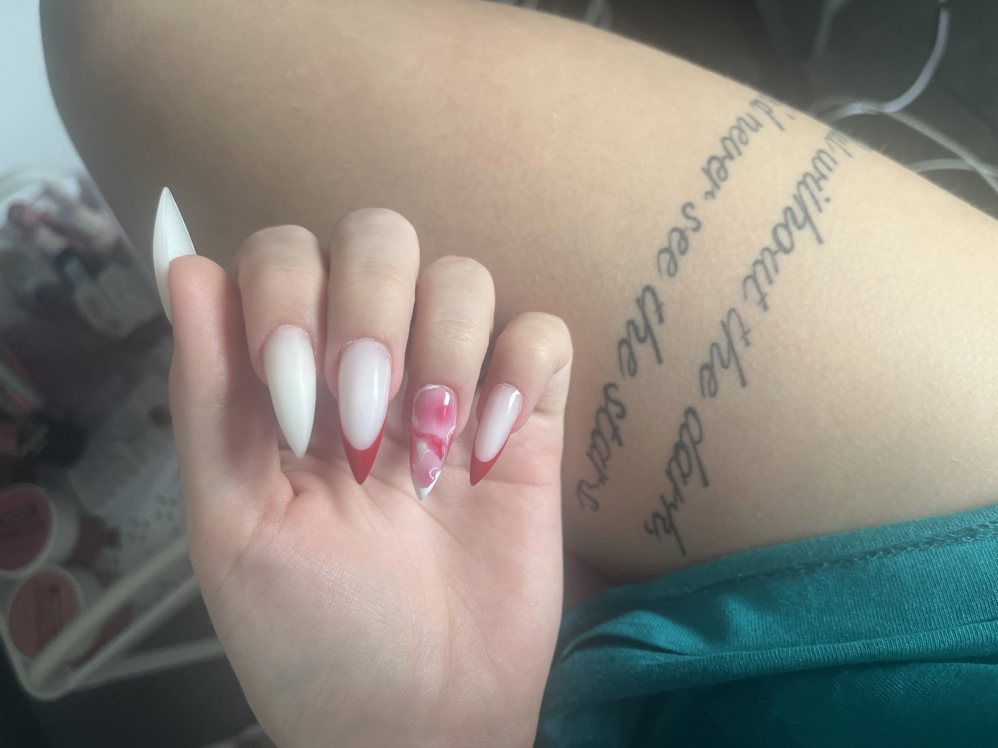 A close-up photograph captures a person's hand with long, pointed nails resting against their leg. The nail on the index finger is entirely white, while the middle and pinky fingers feature a white base with a red French tip. The ring finger nail has a white base with a red and white marble design. The person's hand is fair-skinned. The leg is also fair-skinned and has a tattoo of a quote that reads, "without the dark, never see the stars." The person is wearing a teal t-shirt. Some blurred objects are present to the left of the hand, appearing to be makeup containers. The location is not depicted and is unknown; therefore, Varna, Bulgaria, cannot be confirmed as the city.