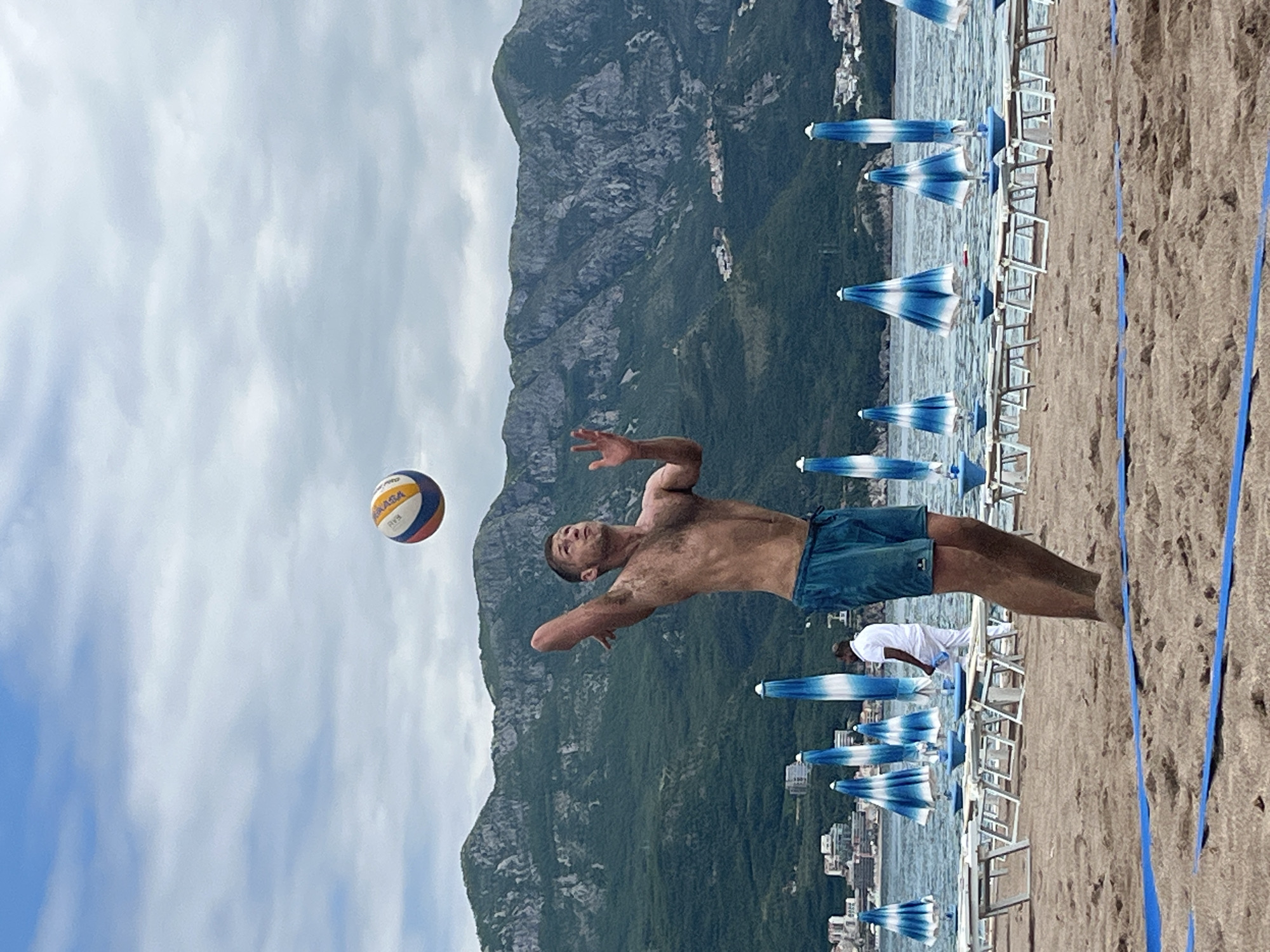 The image shows a beach volleyball game being played in Boreti, Montenegro. A man is diving towards a volleyball in mid-air. Behind him is a mountain range, a beach with blue and white umbrellas, and the city of Boreti. A man in a white shirt is visible near the volleyball player. A blue line marks the boundary of the volleyball court.