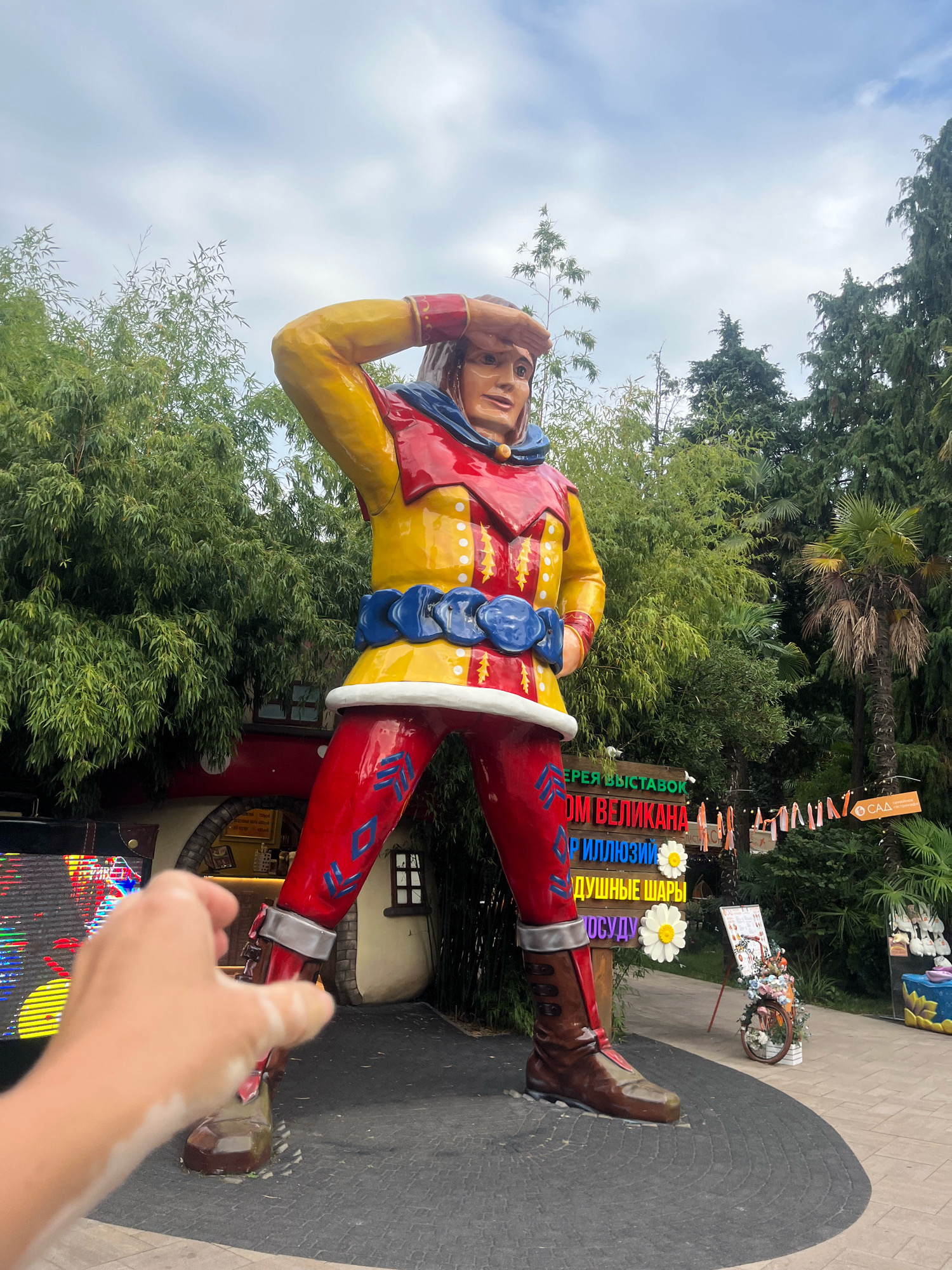 The image shows a large, colorful statue of a man in historical clothing, standing in what appears to be an outdoor recreational area. The statue is dressed in a red and yellow outfit with blue accents and is striking a pose as if shielding his eyes to look into the distance. Behind the statue, there is dense greenery, including bamboo and palm trees, suggesting a warm climate. In the background, there are signs with Russian text and some decorative elements, such as a bicycle adorned with flowers. A person's hand is visible in the foreground, reaching towards the statue, suggesting interaction or a photo opportunity.
This scene is located at **Sochi Park**, an amusement park in the Adler district of Sochi, Russia. The statue itself is likely part of the park's thematic attractions, possibly related to fairy tales or historical figures, given the design and the Russian signage. The park is known for its vibrant decorations, themed areas, and entertainment. The overall atmosphere is one of a cheerful and engaging tourist destination.