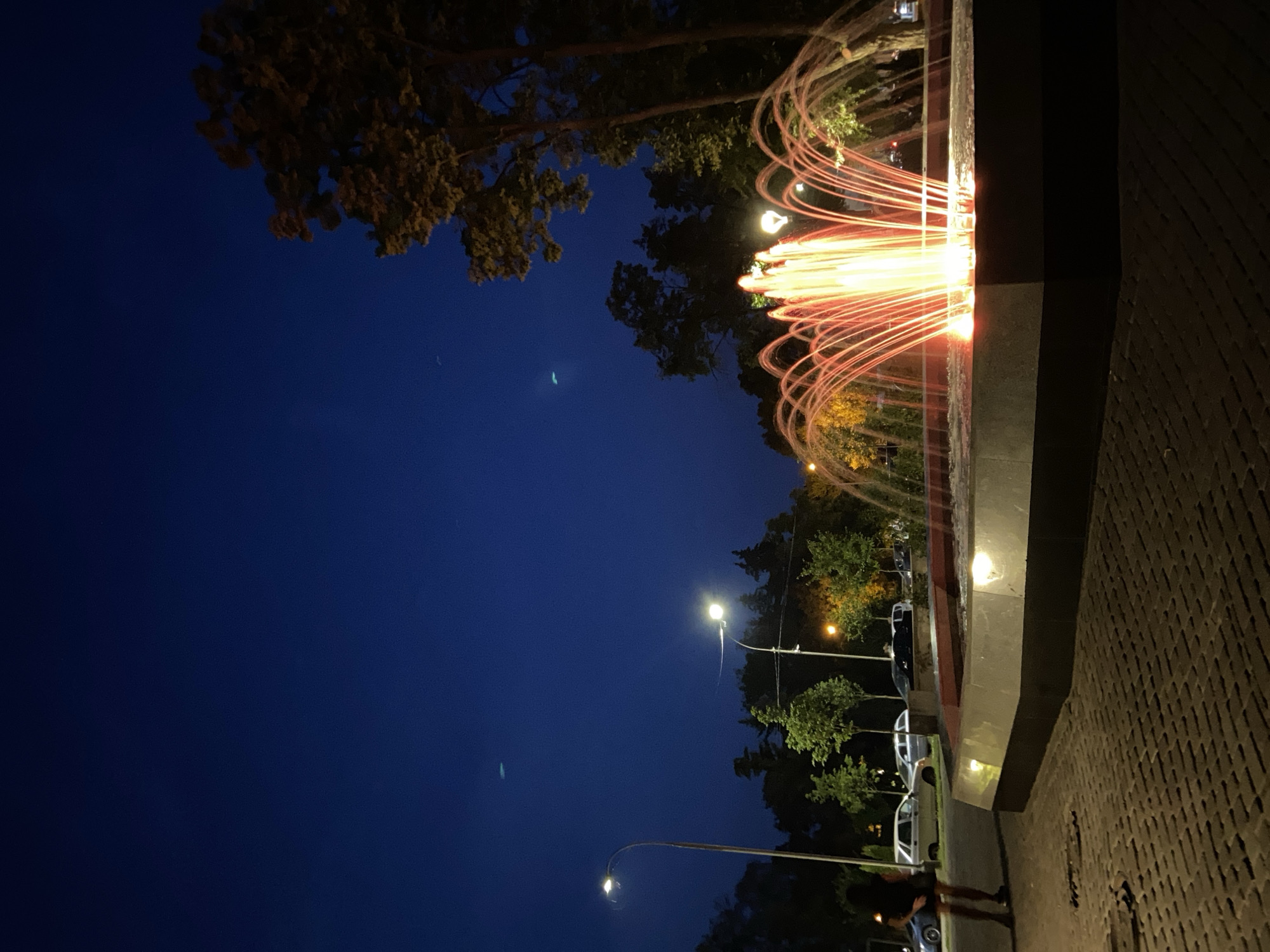 The image depicts a public square at dusk, likely in the city of Poltava, Ukraine. A brightly lit fountain with reddish-orange illuminated water jets is the central focus, situated on a paved area. In the foreground, the paving stones are visible. To the left, a woman stands on the sidewalk, facing away from the camera, with parked cars behind her. Streetlights illuminate the area, casting a warm glow on the surrounding trees and foliage. The sky above is a deep blue, indicating the transition from day to night.