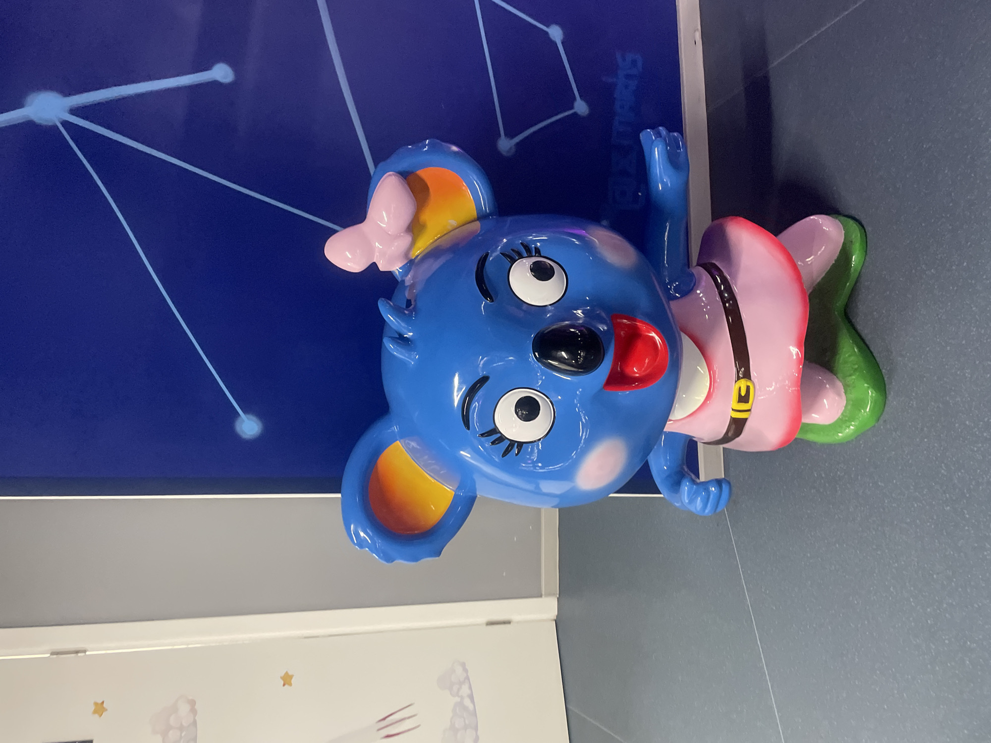 The image depicts a colorful, cartoon-like sculpture of a koala. The koala is primarily blue with yellow accents inside its ears and a pink bow. It is wearing a pink outfit with a belt and a green section at the bottom. The koala's tongue is sticking out, and it has large, expressive eyes. The sculpture is located in Sochi, Russia, as evidenced by the inscription above the statue that reads "Sochi." The sculpture is placed against a backdrop of a dark blue wall adorned with white constellation-like patterns. The surrounding environment consists of a gray wall and the corner of a white panel with drawings on it.
