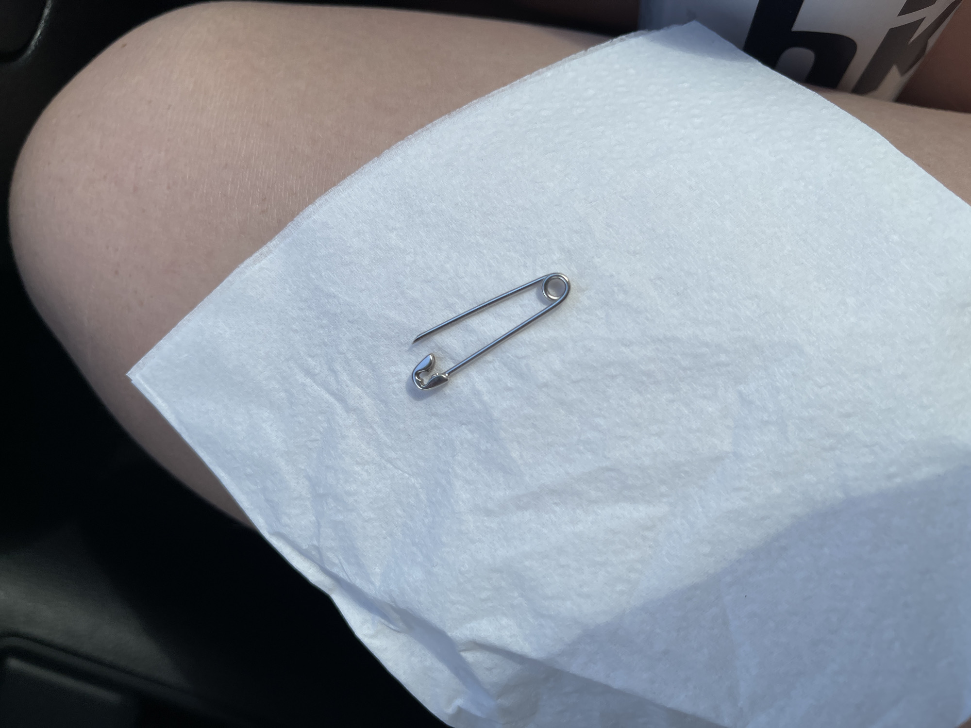A safety pin rests on a white paper napkin, which is placed on a person's leg. The background is dark, suggesting an indoor or shaded environment. There are no specific details to indicate the location in the city of Synhai, Ukraine.