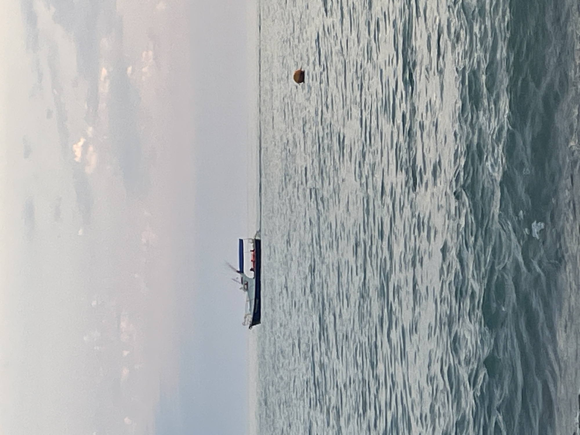 An eye-level, medium shot captures a fishing boat moving across calm, gray water under a hazy, overcast sky. The boat, dark blue and white with an attached blue canopy, has several figures visible on deck. In the middle ground to the right, a single orange buoy bobs on the water. The water's surface is textured with ripples and small waves, particularly in the foreground. The overall scene is serene and devoid of specific identifiable landmarks, suggesting an open body of water.