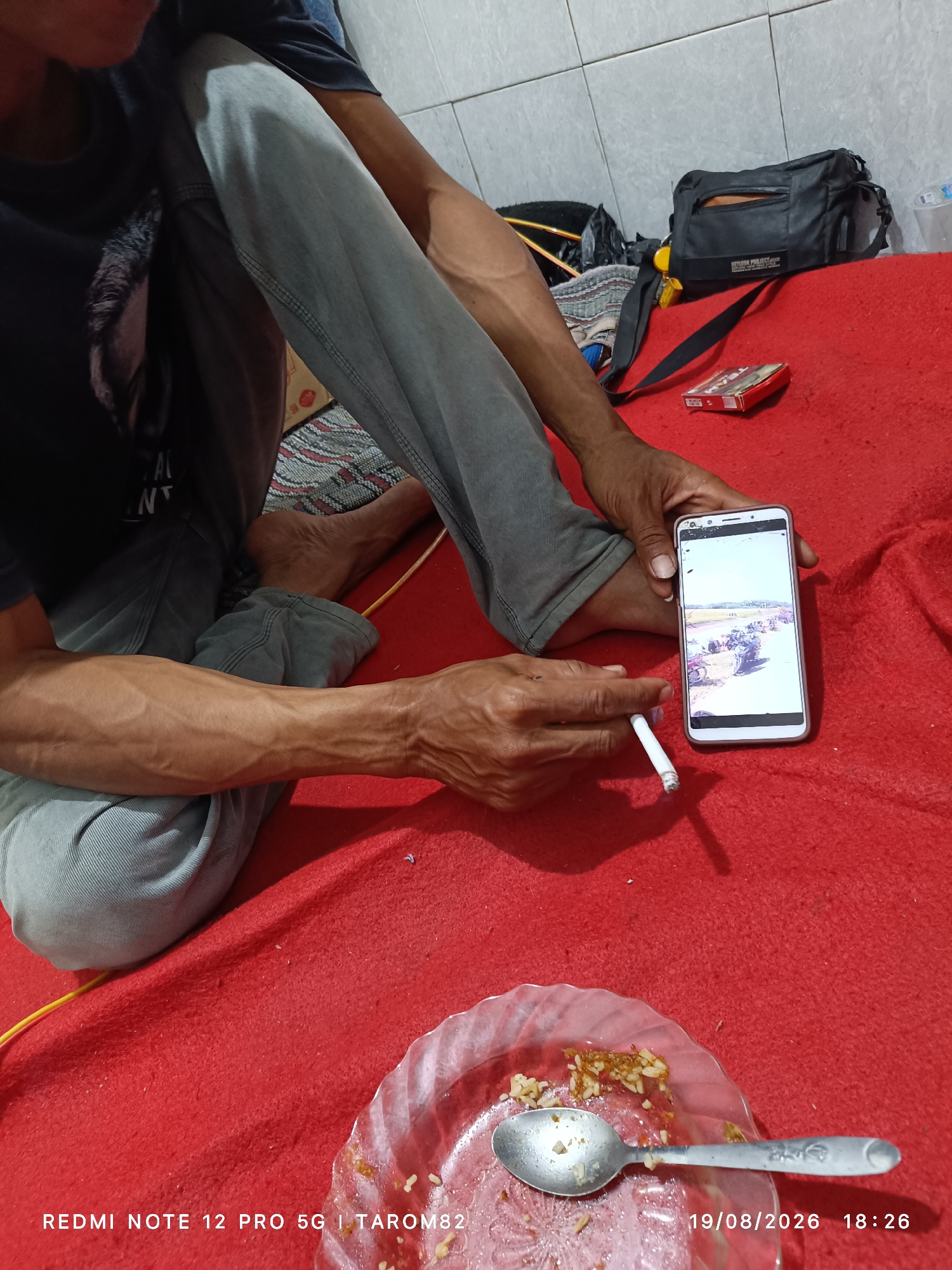 The image depicts a person sitting on a red rug indoors, possibly in the city of Sidodadi, Indonesia. The person is wearing a dark t-shirt and grey pants. They are holding a white cigarette in their right hand and a smartphone in their left hand. The smartphone screen displays a photograph of a line of parked motorcycles outdoors. Nearby, on the red rug, is a cigarette pack and a small black bag. In the foreground, a clear plastic plate with leftover rice and a spoon is visible. The bottom of the image shows a timestamp "19/08/2026 18:26" and text that reads "REDMI NOTE 12 PRO 5G | TAROM82". The environment appears to be a simple room with tiled walls.