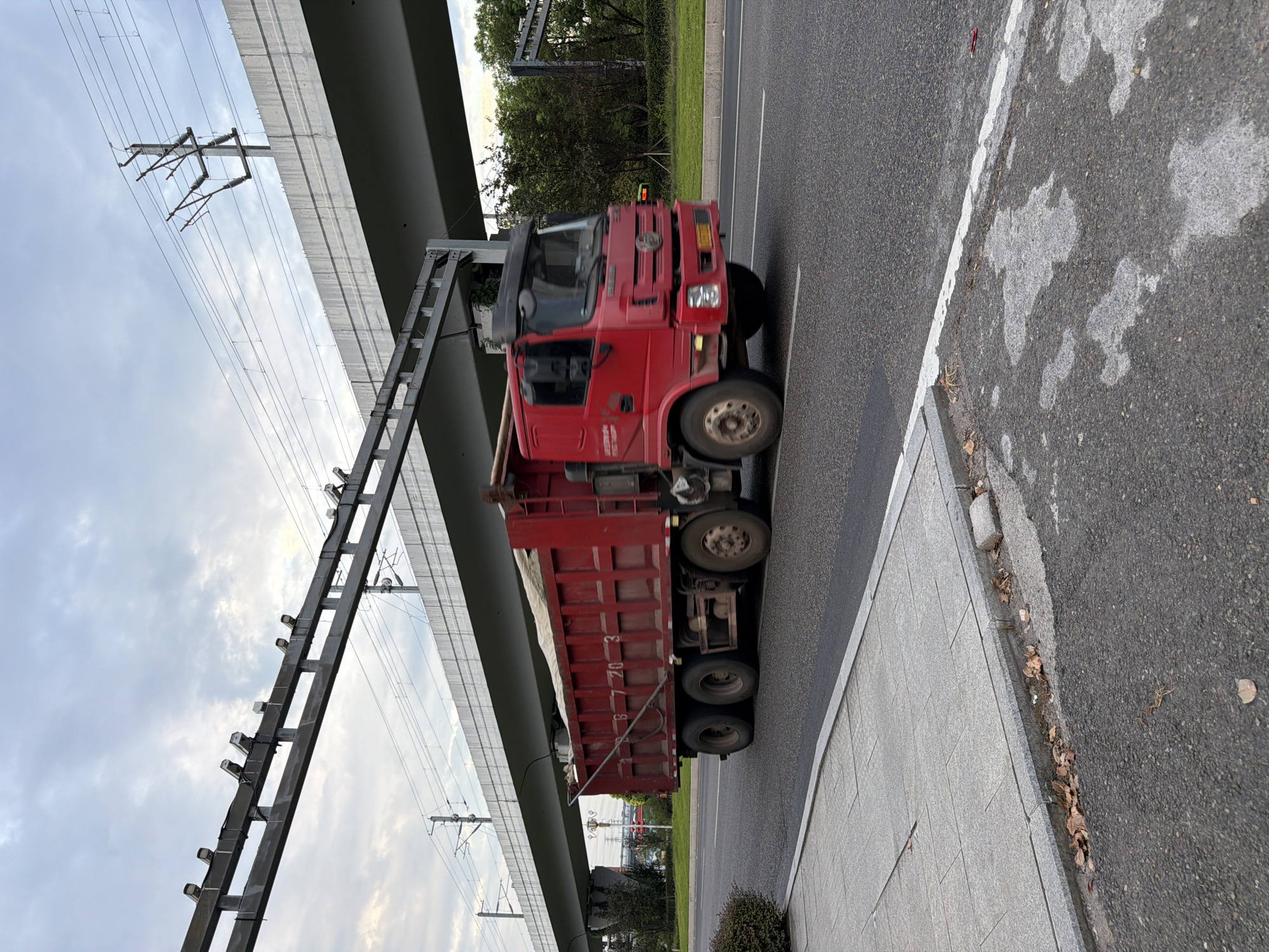 A red dump truck with a load of sand is traveling on a road in Jiangjin District, Chongqing, China. The truck is on the right side of the road and is in motion. The truck has a license plate that reads "B 7 20 3". Overhead, there is an elevated structure, possibly a bridge or overpass, with what appear to be power lines and cameras attached to it. The sky is cloudy. The surrounding environment includes trees, grass, and a sidewalk. The road surface is asphalt with white lane markings.