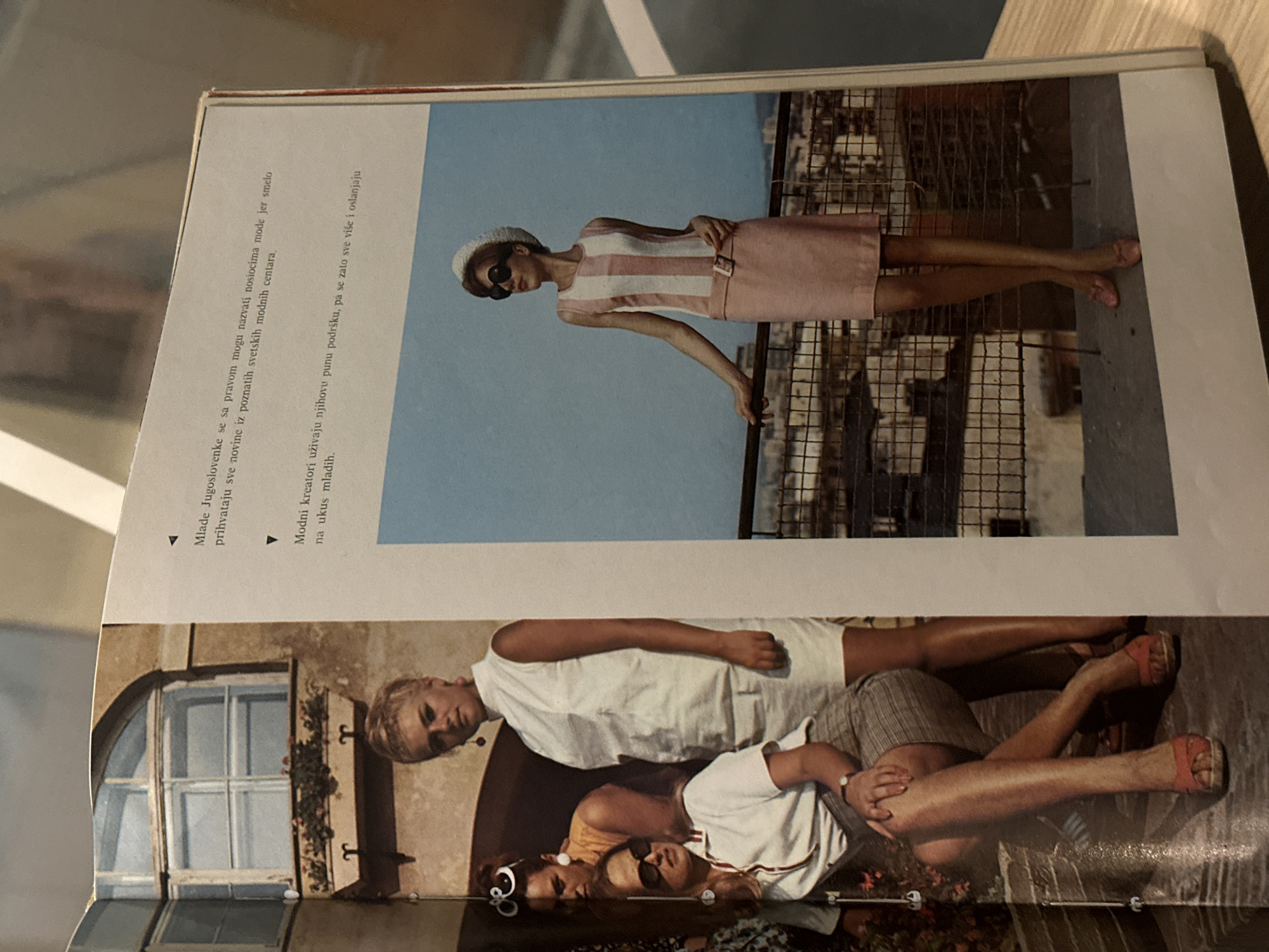 The image shows a magazine spread. The spread features two fashion photos. The upper photo depicts a woman in a pink and white striped dress, sunglasses, and a hat, posing against a metal railing with a blue sky behind her. The lower photo shows three women lying down. The leftmost woman is wearing a white top and sunglasses, with curlers in her hair. Another woman is wearing a white dress, and the third is wearing a white top and striped shorts. The lower photo also includes a building with a window and greenery. The magazine text is in Croatian. The image does not include the location of Novi Sad.