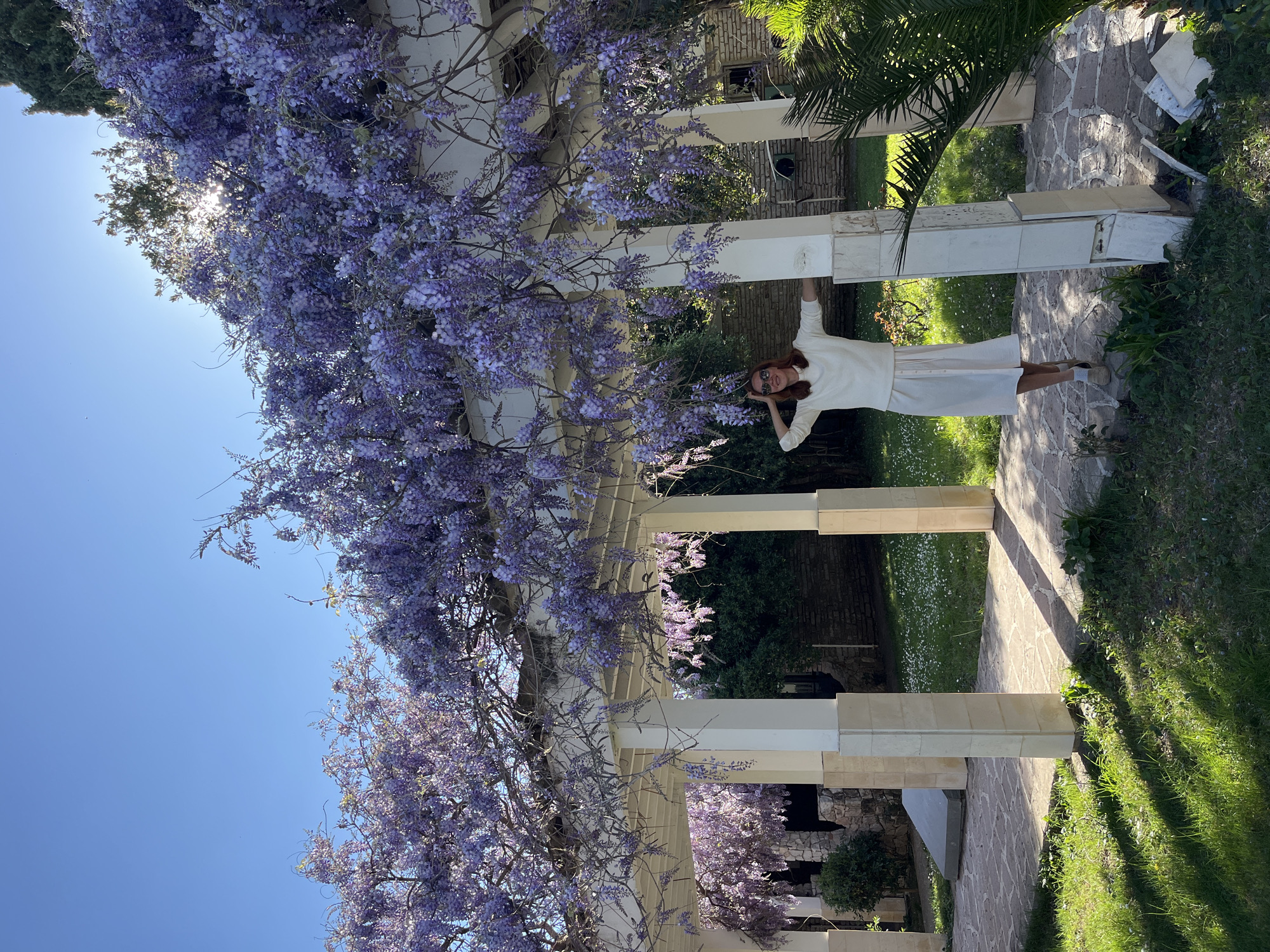 A medium shot depicts a woman standing beneath a wisteria-covered pergola. The woman, fair-skinned and wearing a white outfit, has her hands raised near her head and sunglasses on her face. The pergola structure is constructed of white pillars and horizontal beams. Wisteria blooms profusely along the top of the pergola, with its purple flowers cascading down. The location appears to be a garden or courtyard, with a stone pathway under the pergola and grass and other vegetation on either side. The sky is visible in the upper left corner. A palm tree is visible beyond the right side of the pergola. It is Boreti, Montenegro.