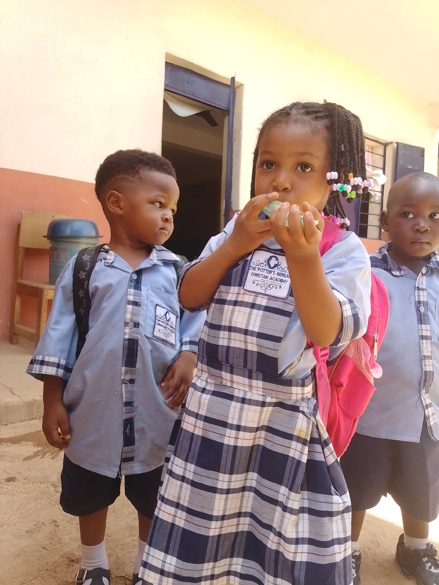 A bright, sunlit outdoor photograph features three young children, likely students, dressed in school uniforms. The setting appears to be a schoolyard or a paved area next to a building with light-colored walls. The ground is a mix of dirt and concrete, suggesting an outdoor space.
In the center, a young girl with black braided hair, adorned with colorful beads (pink, green, orange, white), is prominently featured. She wears a light blue short-sleeved uniform shirt layered under a dark blue and white plaid dress. A vibrant pink backpack is on her back. She is holding a small, light green, translucent object to her mouth, appearing to eat or drink it, and gazes slightly upwards with a thoughtful expression.
To her left stands a young boy with short, dark hair. He wears a light blue short-sleeved shirt with a plaid pattern on the sleeve cuffs and button placket, paired with dark shorts and a black backpack. He looks towards the viewer's left, with a focused gaze. Behind him, part of a wooden chair and a blue plastic drum or bin are visible.
To the right of the girl, another young boy with short hair is partially visible. He is dressed in a similar light blue and plaid uniform shirt and dark shorts. He has a light blue backpack and is also looking towards the viewer's left.
On the left shirt pocket of both the girl and the boy on the far left, a white patch with a logo is visible. The text clearly reads: "THE POTTER'S BEREAN CHRISTIAN ACADEMY". Below this, smaller, less legible text is present, and a globe-like icon is part of the emblem. The bright daylight indicates it is daytime, casting clear shadows, and the overall mood is calm and observational.