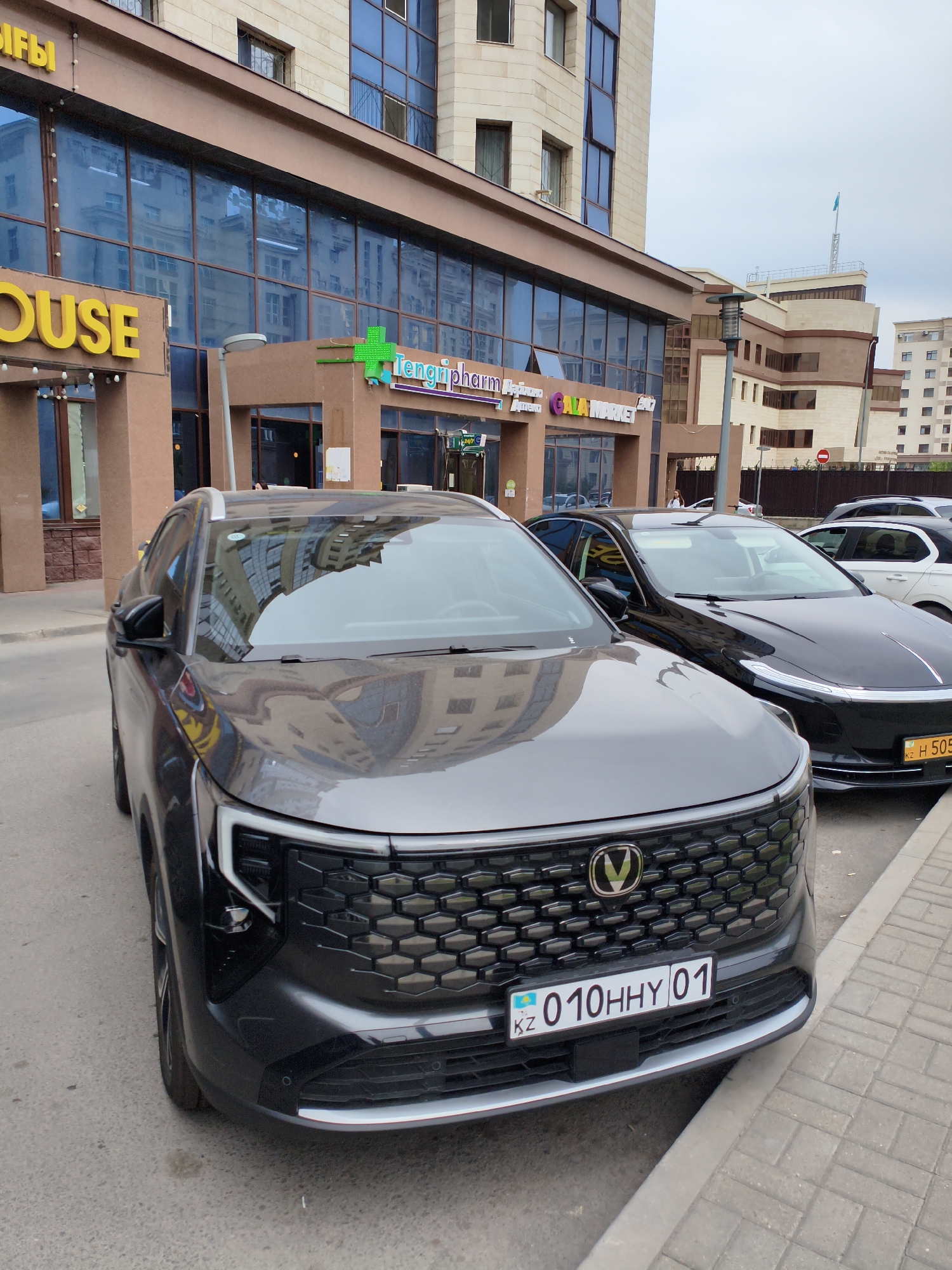The image shows a street scene in Astana, Kazakhstan, with a modern building complex in the background. In the foreground, two cars are parked parallel to the sidewalk. The car closest to the viewer is a dark grey SUV, its front end prominently displayed. It features a distinctive honeycomb-patterned grille with a circular emblem at the top. The license plate of this vehicle reads "KZ 010HHY 01". To the right of the SUV is a black sedan, its front also visible. Its license plate is partially obscured but appears to start with "KZ H 505". The building behind the cars has large glass windows reflecting the sky and surrounding structures. Several signs are visible on the building's facade, including "HOUSE" in yellow lettering and "Tengripharm" with a green cross logo. A street lamp is also visible between the cars. The sidewalk is paved with brick-like stones. The sky is overcast.