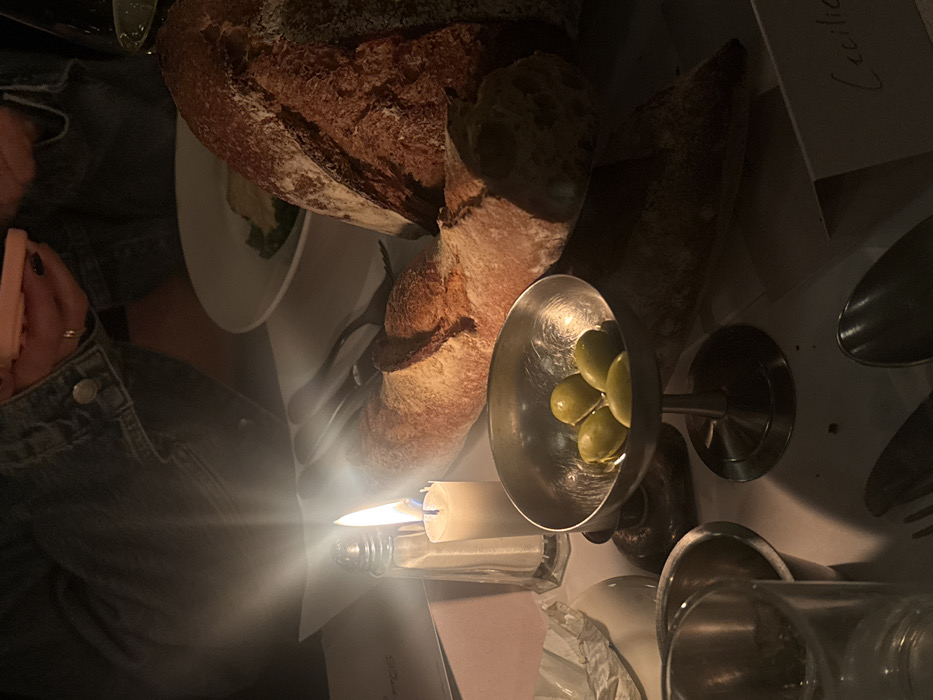 A table setting in dim light features bread, olives in a silver dish, cutlery, and a lit candle. A partially visible person in a denim jacket holds a pink phone. A plate of greens is also present. The setting suggests a restaurant or dining experience.