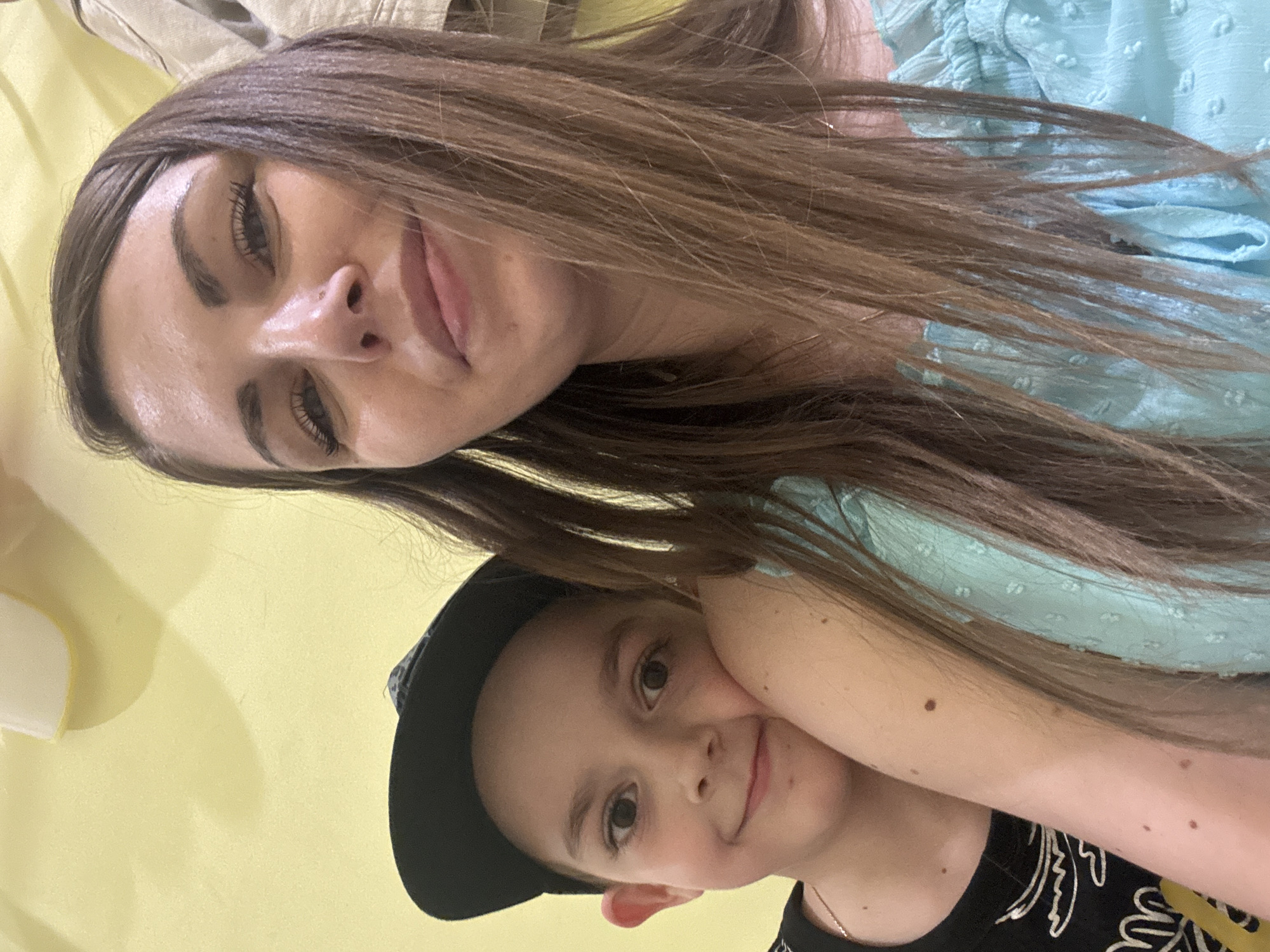 A selfie-style image features a woman and a young boy. The woman has long brown hair, fair skin, and is wearing a light blue top. She appears to be holding the camera. The boy, wearing a black baseball cap and a black shirt with a white pattern, is positioned below her. The background is a pale yellow wall. There is no discernible evidence to identify a location, or that the image was taken in Shevlyakovo, Russia.