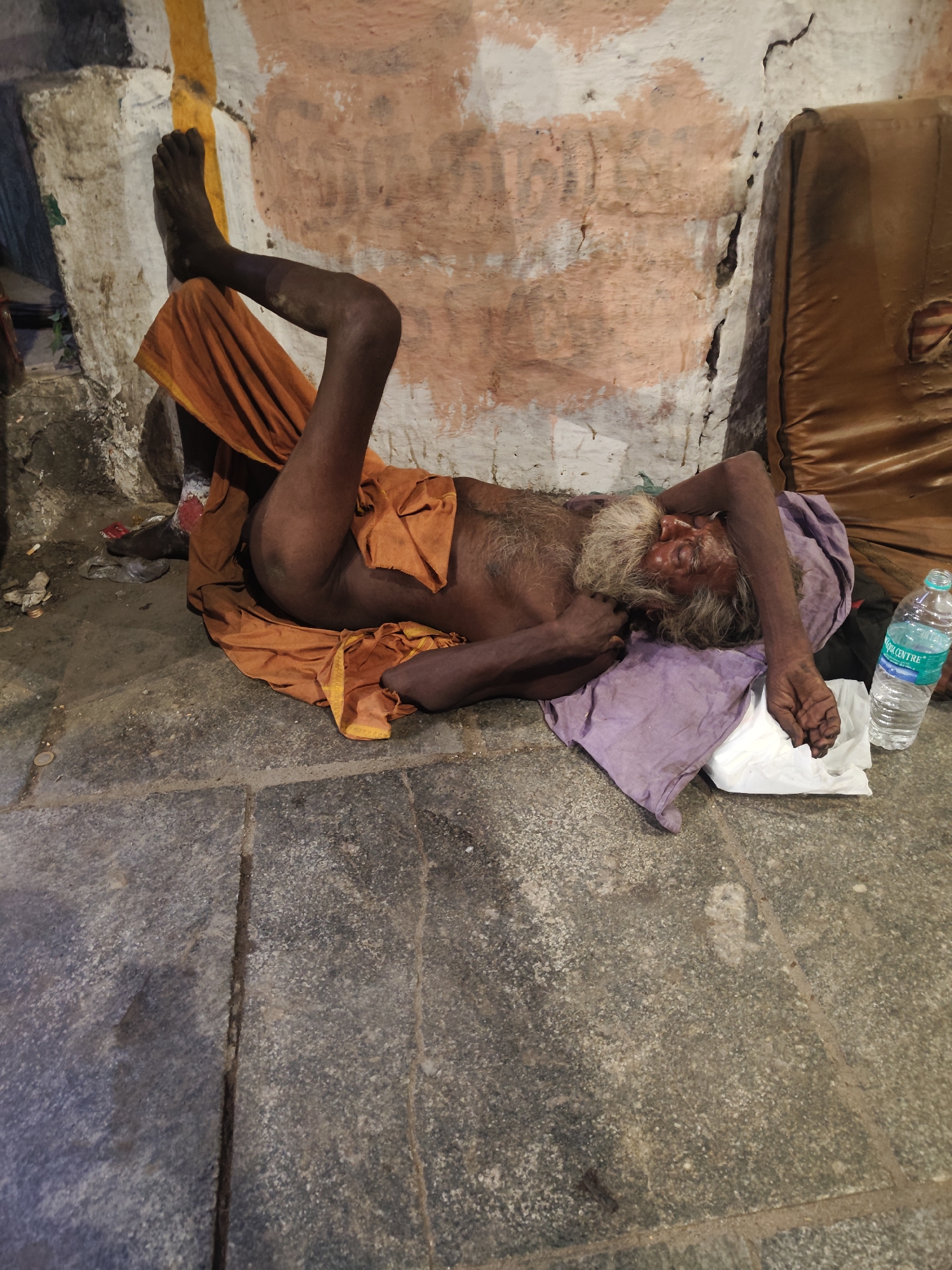 An elderly man is depicted lying on a stone floor, seemingly resting or asleep, within a sheltered area in Chennai, India. He lies on his right side, with his left leg bent and raised against a wall, while his right leg is extended. He is partially covered by an orange cloth wrapped around his lower body, with another orange cloth draped near his left hip. His upper torso is bare.
The man has a long, prominent white and grey beard and sparse hair. His face, seen in profile, appears aged, and his eyes are closed. His head rests on a crumpled purple cloth or pillow. His right arm is raised and extended above his head, while his left arm is bent with his hand near his chest. Adjacent to his head, a white plastic bag and a clear plastic water bottle are visible. The water bottle's label clearly displays the text "AQUA CENTRE."
The floor is composed of large, dark grey stone slabs with discernible grout lines. The wall behind the man is weathered and textured, featuring patches of faded white and peachy-orange paint, along with a prominent vertical yellow stripe. The wall shows signs of cracking and wear. To the man's right, a brown, padded object, possibly a worn mattress or cushion, leans against the wall. A closer inspection of the man's left foot reveals a red mark, possibly a religious tilak or kumkum, near the ankle. The diffused lighting suggests an indoor or shaded outdoor setting. The scene reflects a common sight in the region, characteristic of public or semi-public spaces like temple complexes or covered walkways.