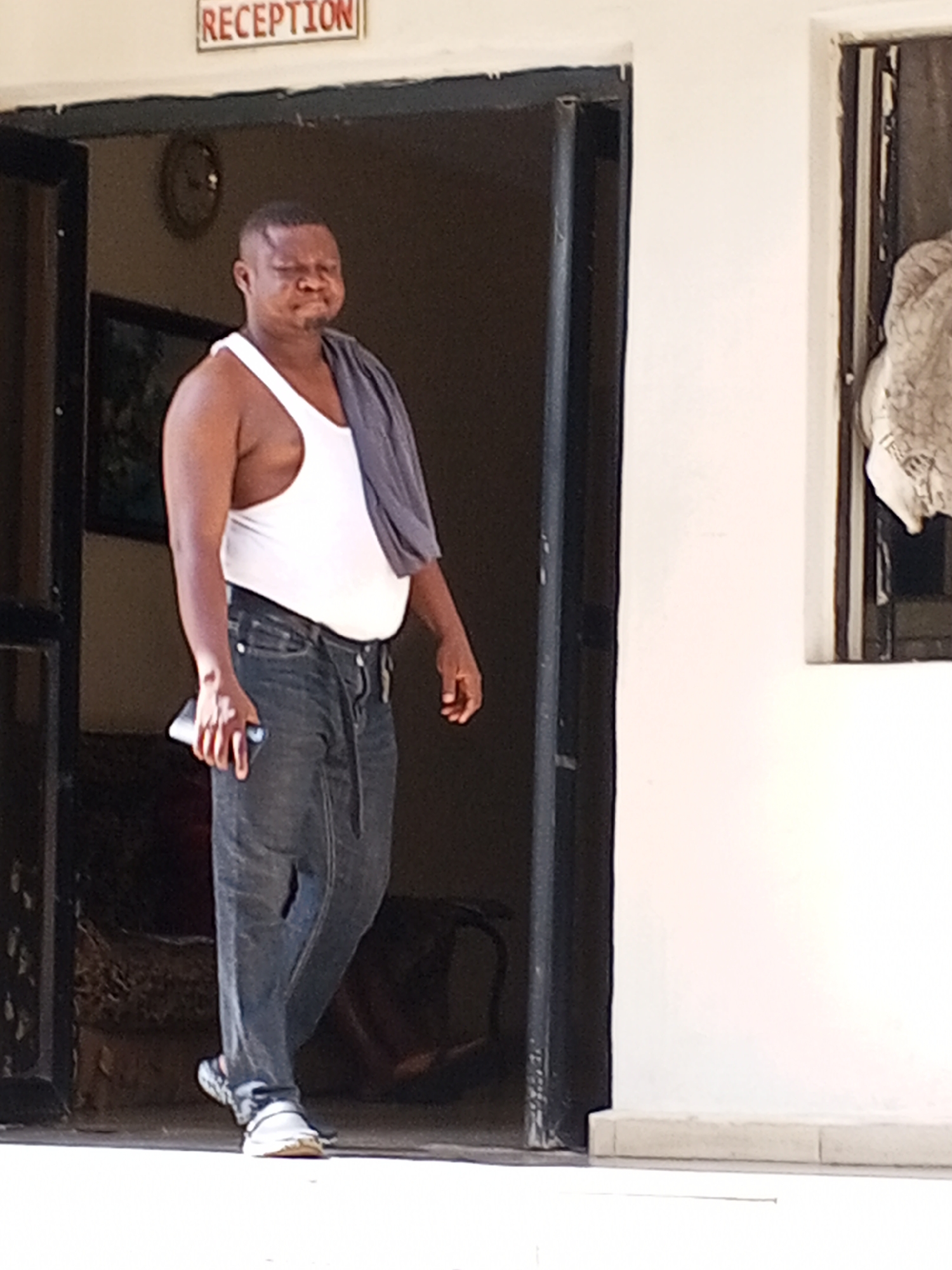 A medium shot shows an adult man walking out of a doorway on a bright day. He has dark skin, is wearing a white tank top, dark blue jeans, and light-colored sneakers. A dark grey cloth is draped over his left shoulder. He appears to have a stocky build. His facial expression is somewhat ambiguous, with eyes squinted, possibly due to the bright light, and cheeks puffed out slightly. He holds a dark-colored smartphone in his left hand.
The man is emerging from a dark-framed doorway. Through the doorway, a dimly lit interior space is visible, containing what looks like a sofa or chair and a circular clock on the back wall. Directly above the doorway, a white sign with red lettering clearly reads "RECEPTION." To the right of the doorway is a plain, light-colored wall, featuring a window or opening with some light-colored, possibly patterned fabric hanging from it. The overall impression is of a casual exit from a building, likely a reception area or lobby, during daylight hours.