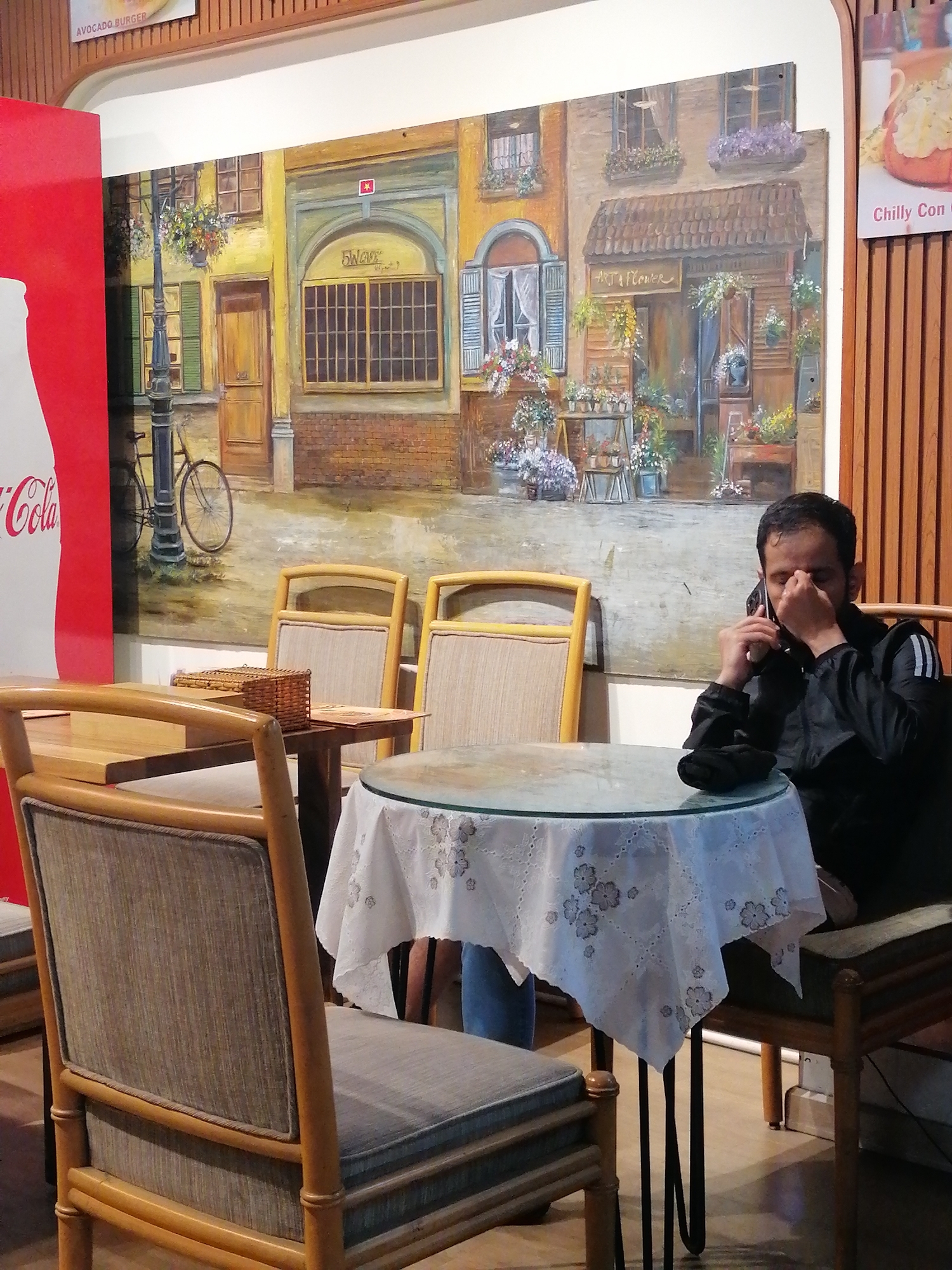 A man, appearing to be of South Asian descent and wearing a black jacket with white stripes on the sleeves, is seated at a small round table covered with a white lace tablecloth. He is speaking on a dark mobile phone, holding it to his right ear with his right hand while his left hand is near his face. The table has a glass top and three legs. In front of him is another wooden chair with upholstered seating and back. To the left of this chair, another similar chair is partially visible.
Behind the man, a large, vibrant mural depicts a European street scene with buildings, windows, flower boxes, a lamppost, and a bicycle. A large red Coca-Cola advertisement is situated to the far left of the mural, partially obscuring it. The man is seated in what appears to be an indoor cafe or restaurant. The flooring is wooden.
The overall atmosphere is casual, with the focus on the man engaged in a phone call. The mural provides a colorful and detailed backdrop. There are no visible contextual clues within the image to specifically indicate the location as Da Lat, Vietnam; however, it is possible this cafe is located there.
