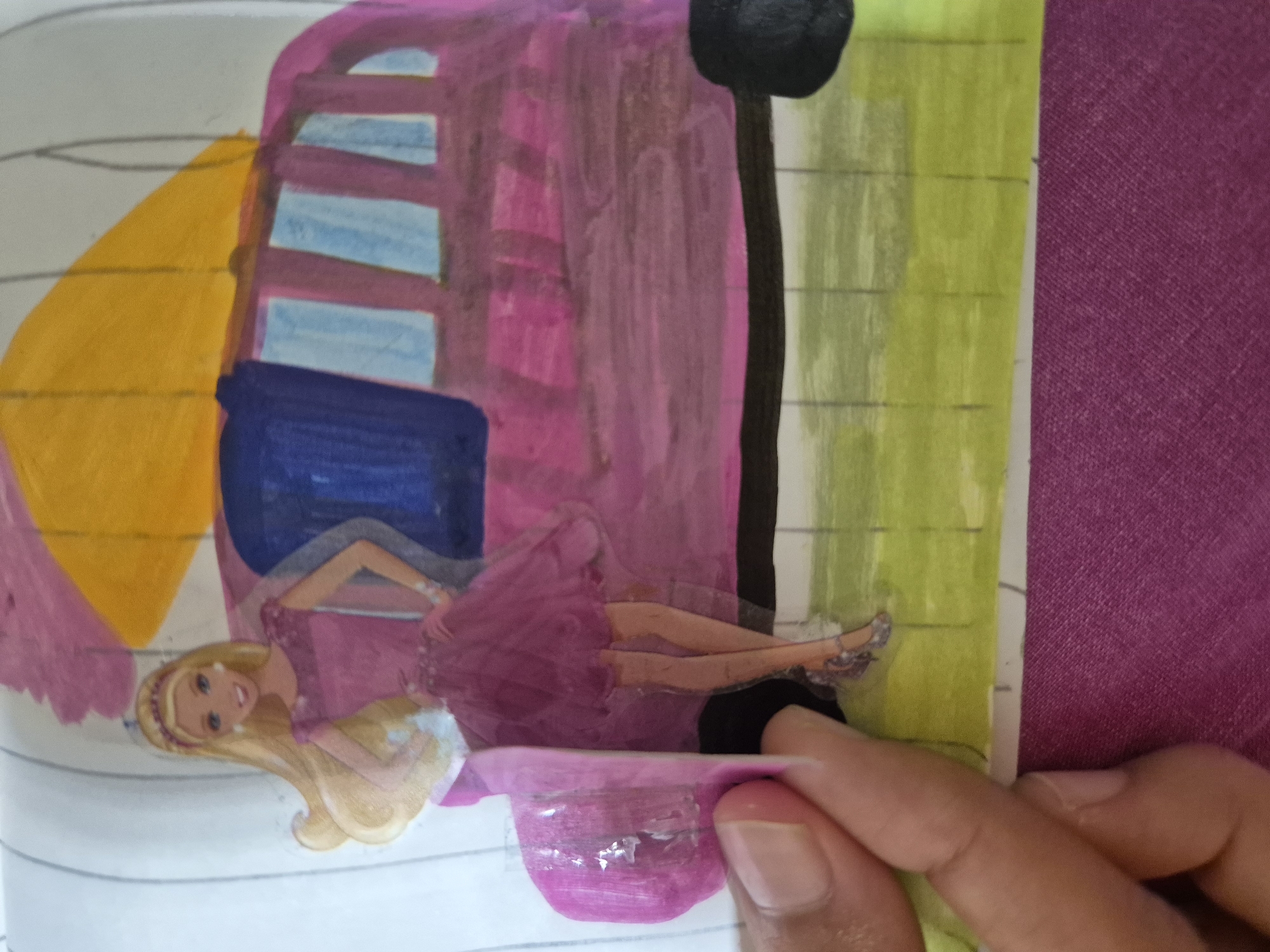 The image shows a close-up of a drawing of a Barbie doll in a pink dress standing in front of a bus. The background is colorful, with a yellow roof and blue windows on the bus. The drawing appears to be made with crayons or colored pencils. A person's hand is visible in the lower left corner of the image, holding up a piece of the drawing. The drawing is on a white piece of paper, which is taped or glued onto a larger surface that appears to be a notebook with a purple cover. The drawing is in a state of disarray, as if it is being handled or modified. There is no indication of the location in the city of Kokshetau, Kazakhstan, or any specific context about the drawing or its purpose.