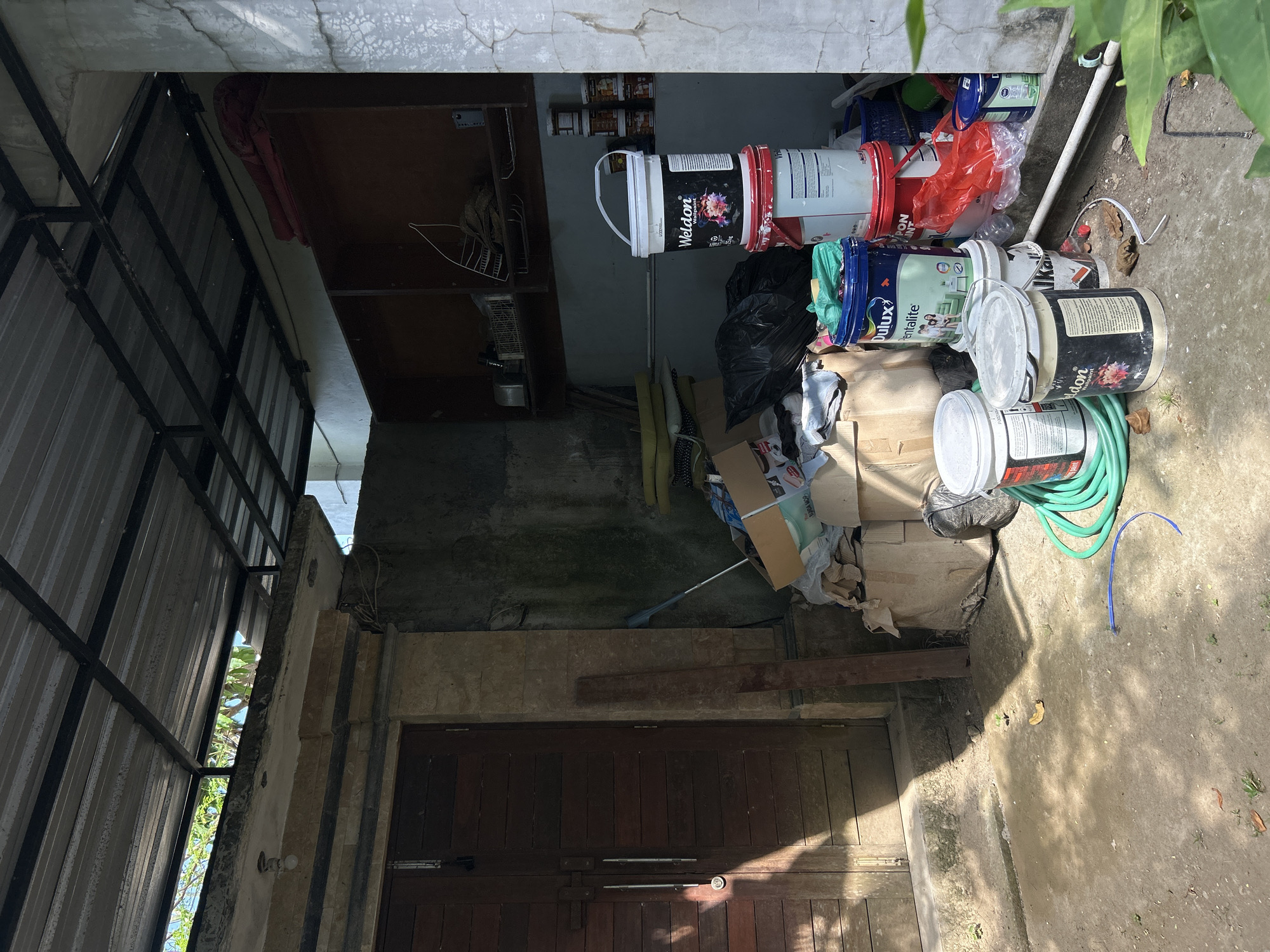 The image depicts an outdoor storage area adjacent to a building, likely a residence or workshop, in North Kuta, Indonesia. The scene is cluttered with various items, suggesting a work-in-progress or a storage space for materials.
Dominating the foreground are several large buckets of paint, stacked and leaning against a wall. One prominent bucket, labeled "Weldon Wallpaper Paint," is white with a black label featuring a floral image. Next to it is a blue bucket labeled "Dulux Pentalite." Other buckets, some red and some white, are also visible, along with their contents and labels, though some are partially obscured. A coiled green garden hose lies on the ground, connecting to a blue hose nozzle.
To the left of the paint buckets, a large wooden door with horizontal planks and silver handles is visible. Sunlight casts a strong shadow across the lower portion of the door and the ground in front of it. Leaning against the wall near the door is a long, slender piece of wood.
In the background, a series of shelves are built into an alcove, containing various items including some red folded fabric or material, what appears to be a wire basket, and smaller paint cans or containers stacked on a shelf. A dark plastic trash bag is also present among the clutter. The walls appear to be concrete, with some exhibiting signs of wear and cracking. The roof above is corrugated metal, supported by a metal frame.
The ground is a mixture of dirt and concrete, with scattered dry leaves. The environment appears somewhat unkempt, with evidence of construction or painting materials and general storage. No individuals are present in the scene. The overall impression is one of a functional but untidy outdoor storage area.