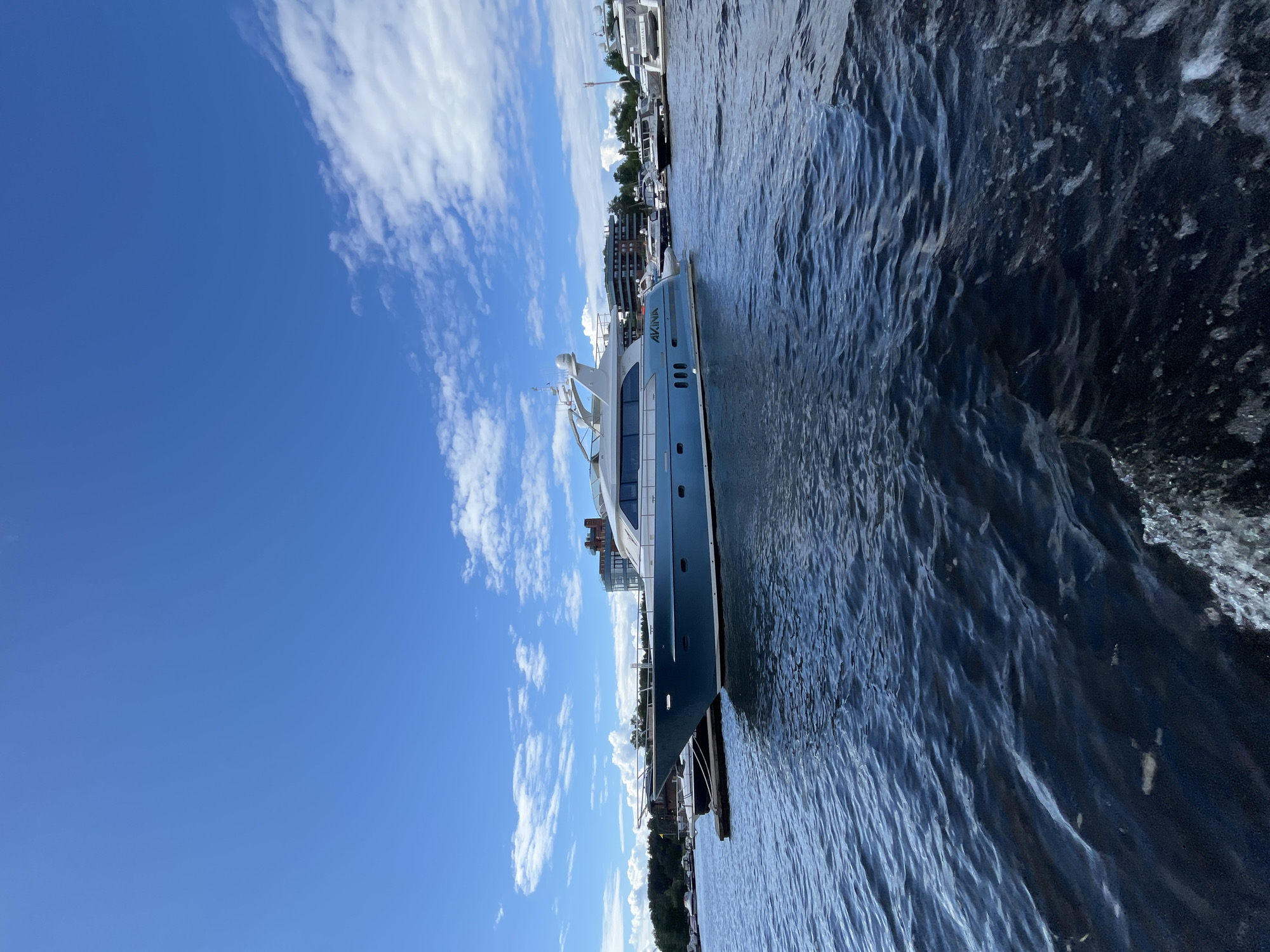 The image captures a serene scene at a marina in Saint Petersburg, Russia. A luxurious yacht, named "AVALON" as indicated by text on its hull, is docked and takes center stage. The yacht is sleek and modern, painted in a striking blue and white color scheme, with large windows suggesting a comfortable interior.
The yacht is moored alongside other vessels and a waterfront promenade. In the background, buildings of varying architectural styles are visible, hinting at the urban landscape of Saint Petersburg. Trees line the shore, adding a touch of nature to the scene.
The sky is a vibrant blue, dotted with fluffy white clouds, suggesting fair weather and a pleasant day. The water is calm with gentle ripples, reflecting the blue sky and the surrounding boats. The perspective is from a low angle, looking across the water towards the yacht. The time of day appears to be daytime, likely late morning or early afternoon, given the bright sunlight and the quality of the light. There are no people visible in the frame, creating a sense of peaceful solitude. The overall mood is tranquil and picturesque, showcasing a moment of calm amidst a bustling port.