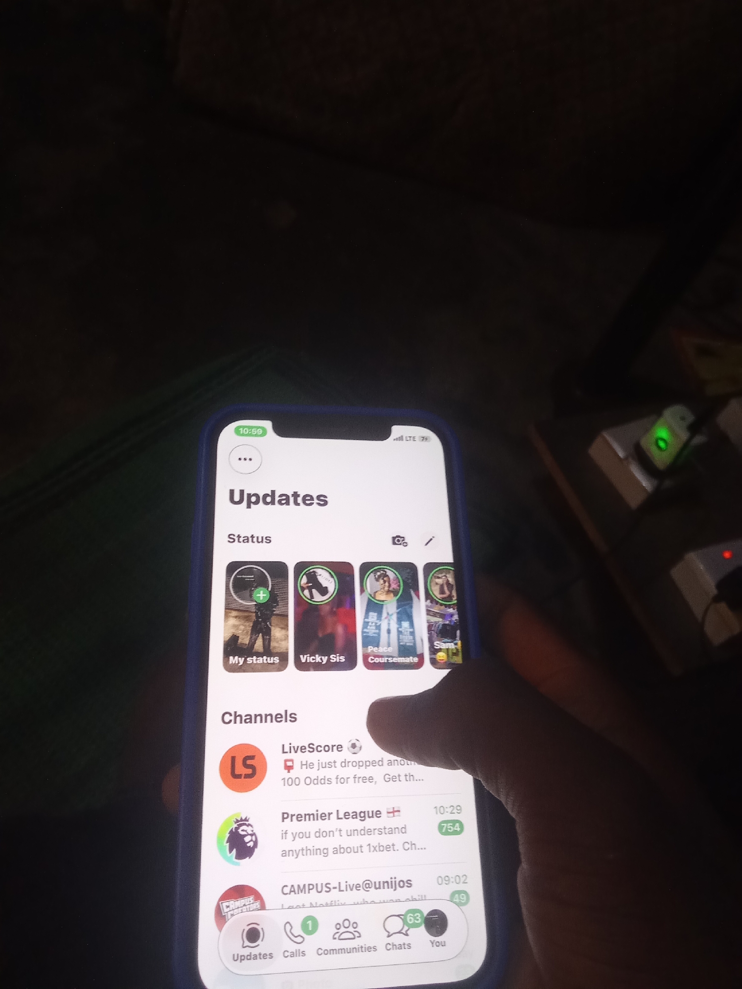The image shows a close-up of a smartphone screen displaying a social media application, likely WhatsApp or a similar messaging platform, with the "Updates" tab selected. The screen is oriented vertically.
At the top of the screen, the time is displayed as "10:59". Below this, the header "Updates" is visible, along with a three-dot icon and a camera icon, suggesting options for creating or viewing content.
The "Status" section features several circular profile pictures, including "My status" with a green plus icon, "Vicky Sis", "Course mate", and "Sam". These likely represent user-created status updates.
The "Channels" section lists several channels. The first is "LiveScore" with a soccer ball icon and a message mentioning "100 Odds for free". The second is "Premier League" with its official logo and a message about understanding "1xbet". The third is "CAMPUS-Live@unijos" with a message about "Netflix".
At the bottom of the screen, there is a navigation bar with icons for "Updates", "Calls", "Communities", "Chats", and "You". The "Updates" icon is highlighted.
The smartphone is being held by a person whose hand is partially visible on the right side of the screen, obscuring some of the phone and the surrounding environment. The background is dark and out of focus, with some electrical outlets and a green LED light visible in the upper right corner, suggesting an indoor setting, possibly a room with electronic devices. The overall lighting is dim, with the phone screen being the primary light source. The location is not specified and cannot be determined from the image.