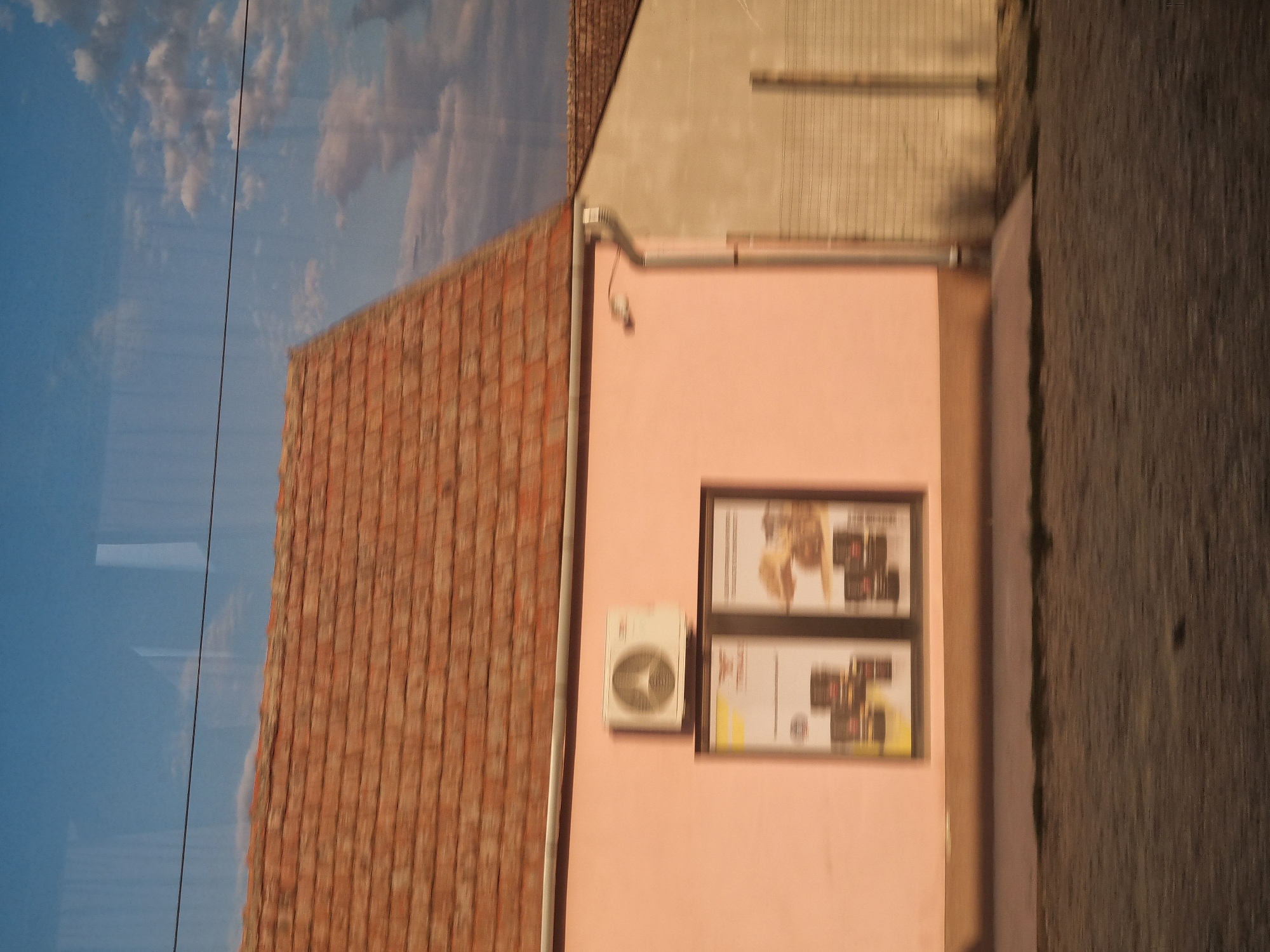 The image depicts a building with a pink facade and a red tile roof, likely located in the city of Nojorid, Romania. An air conditioning unit is mounted on the pink wall, above two framed displays that show images of products. The building is adjacent to another structure with a beige facade and a similar tiled roof. A wire runs horizontally across the sky, and clouds are visible in the blue sky. The ground in front of the buildings appears to be a paved area. The scene is captured with a slight blur, suggesting movement.
