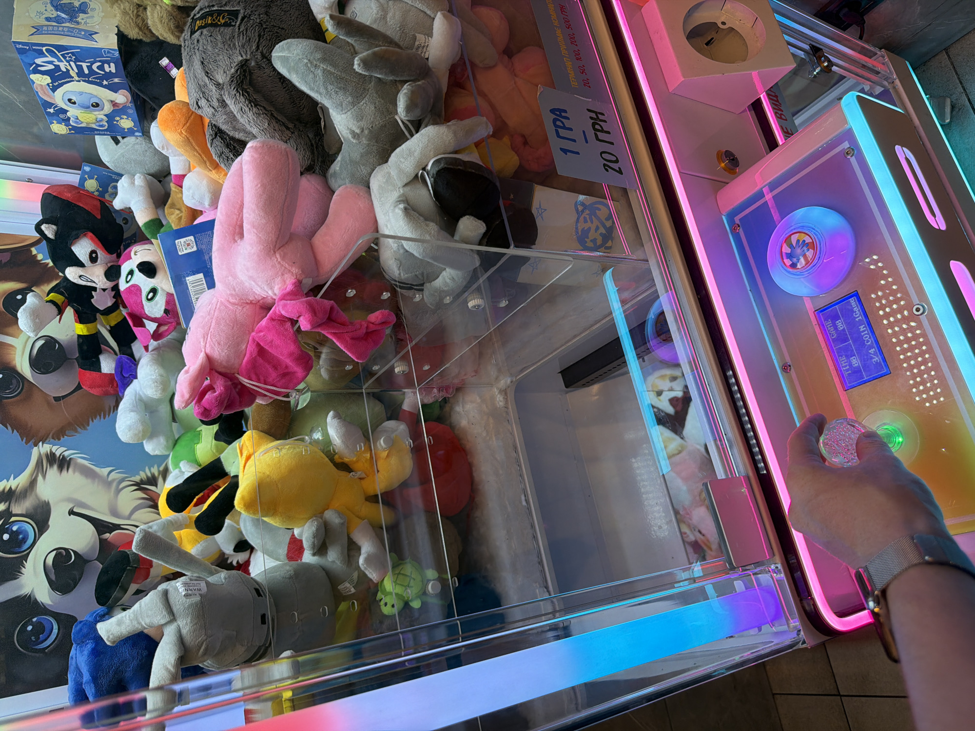 In a brightly lit arcade, a person's hand with a silver watch grips a joystick with a colorful, illuminated top. The joystick is positioned on a control panel with a digital display showing "TIME 00:00 GAME 00 3/4 COIN 1GAME". The panel is bordered by vibrant pink and blue LED lights. Behind the panel, a claw machine is filled with a variety of plush toys, including a pink axolotl, a yellow duck, a green turtle, and various animal characters. A price sign in Ukrainian currency indicates "1 GRA 20 GRN". The background features cartoonish dog graphics. This scene is likely located in a public entertainment venue in Kyiv, Ukraine.