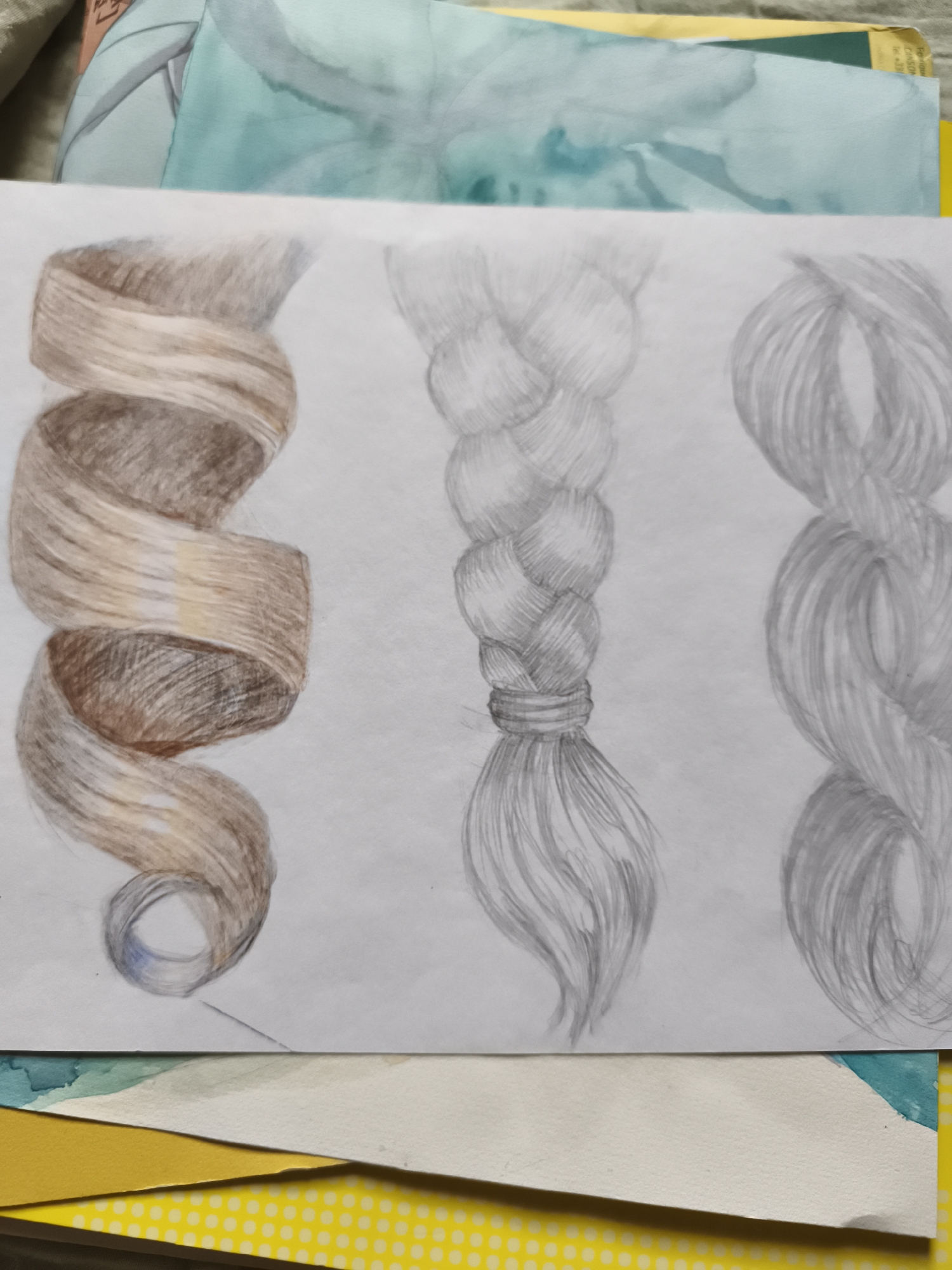 The image displays a drawing of three different hairstyles on a piece of white paper, which is placed on top of other papers and a yellow surface with a white polka dot pattern. The drawing is presented in a close-up view. On the left is a coiled strand of hair rendered in shades of brown and blonde, with visible shading and highlights suggesting texture and volume. In the center, a braided strand of hair is depicted in pencil, with detailed rendering of the weaving and a band tied at the bottom, from which individual strands of hair fall. To the right, another braided strand of hair is drawn in pencil, but less detailed than the central braid, with flowing lines indicating the strands. The drawing appears to be a study of hair textures and styles. The background elements suggest an art or craft setting, possibly an artist's workspace. There is no indication of a specific location within the city of Saint-Denis, France.