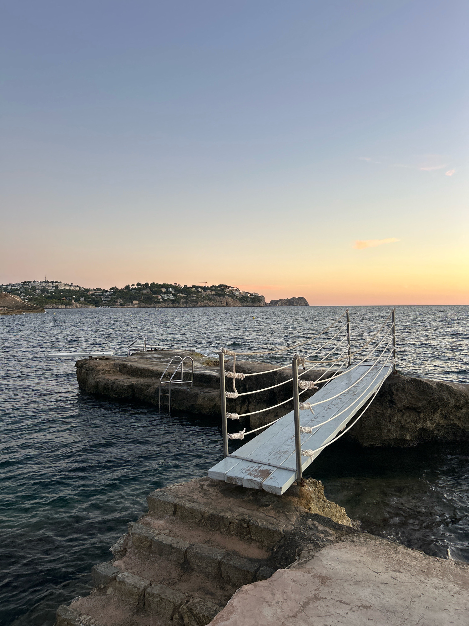 An outdoor shot captures a picturesque coastal scene at dusk in Costa de la Calma, Spain. The ocean, a deep blue, gently laps against rocky formations that jut out from the shore. A weathered stone staircase leads down to the water's edge from a concrete platform. Perched on the rocks are two metal ladders, partially submerged in the sea, suggesting a swimming area. A narrow wooden walkway, suspended by ropes on either side, extends from one rock to another, leading towards the open water. In the background, a hillside is dotted with houses, illuminated by the soft glow of the setting sun. The sky transitions from a pale blue overhead to a warm orange and pink hue near the horizon, where a faint cloud drifts. The overall impression is one of serene natural beauty and a well-loved spot for seaside relaxation.
