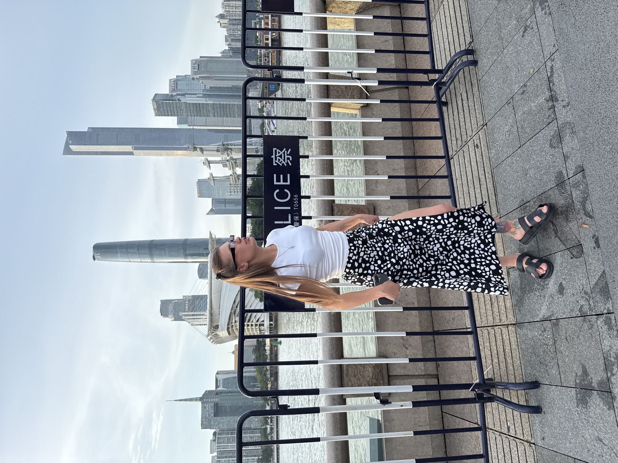 A young woman with long blonde hair, wearing glasses, a white t-shirt, and a black and white polka-dot skirt stands on a paved walkway. She is looking to her right. Behind her, a body of water is visible, and beyond that, a cityscape with numerous tall buildings. A black and white striped barrier runs across the middle ground, separating the walkway from the water. A sign on the barrier reads "POLICE" in English and Chinese characters. The scene appears to be in Haizhu District, China, likely along the Pearl River. The woman is holding a dark-colored smartphone in her right hand. She is wearing black sandals, and a tattoo is visible on her left calf. The sky is overcast with some clouds.