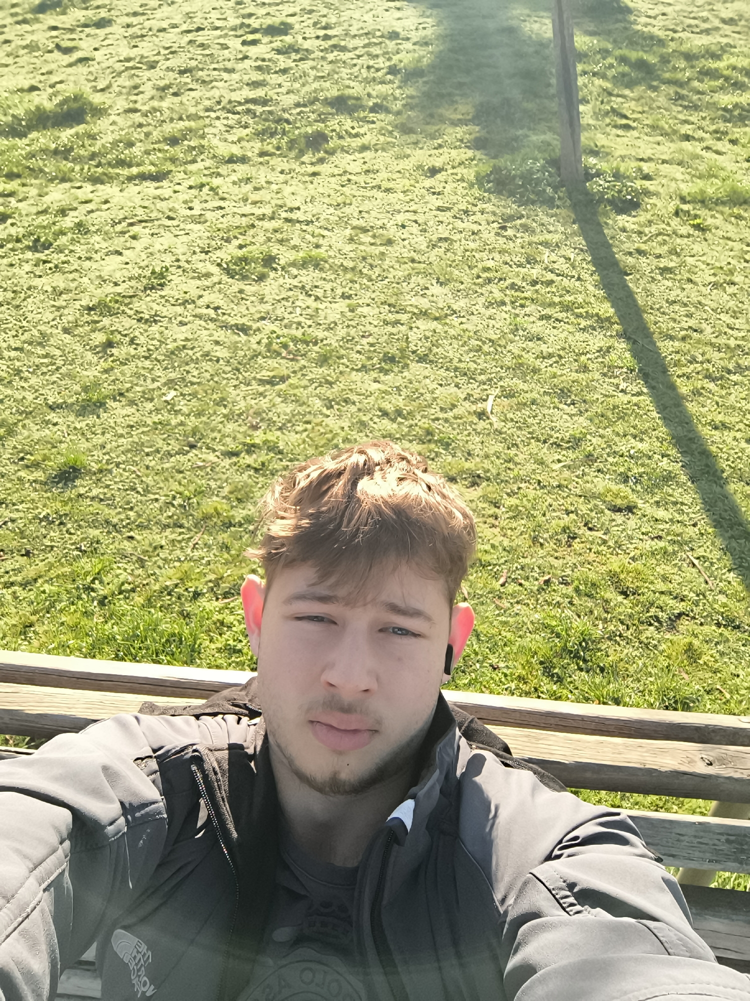 A selfie taken by a young adult male with light brown hair and blue eyes, wearing a dark gray jacket and a black earbud in his left ear. He is sitting on a wooden park bench. The background consists of a green grassy area, potentially a park. A thin tree trunk and its shadow are visible in the upper right. The image was likely taken in an outdoor, possibly sunny environment. It is not possible to determine if this image was taken in Sofia, Bulgaria based on only the given image.