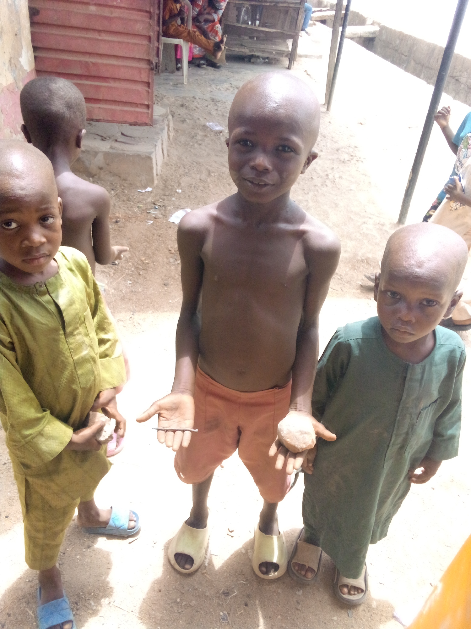 Three young boys stand outdoors in Minna, Nigeria. The central boy, shirtless and wearing orange shorts and cream-colored sandals, holds a nail in one hand and a small rock in the other. To his left, a boy in a light green long-sleeved garment and blue sandals holds an unidentifiable object. To his right, a boy in a dark green long-sleeved garment and brown sandals stands next to the central boy. A fourth boy stands shirtless in the background, his back to the camera. The setting is a dirt area near a building with a red door. Other buildings and people can be seen in the background.