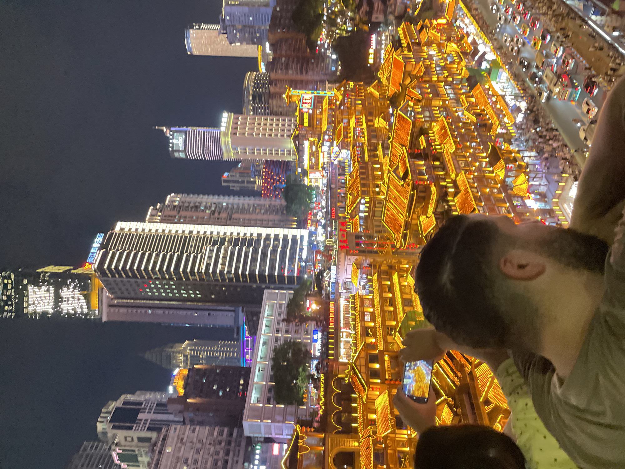 The image is a nighttime aerial view of a densely populated urban area, likely Chongqing's Yuzhong District in China, characterized by towering skyscrapers and a vibrant, multi-story traditional building complex illuminated with warm, golden lights. In the foreground, two individuals are visible, with the shoulder and back of one person prominently featured. This person appears to be looking over the cityscape. Another person's head is partially visible, and they are holding a smartphone to take a picture of the illuminated buildings. The background showcases a dense urban environment with various modern skyscrapers, some adorned with light displays. Below the traditional buildings, a street with traffic, including cars and a bus, is visible, along with a significant number of pedestrians. The overall impression is of a bustling city at night, blending modern architecture with traditional design.