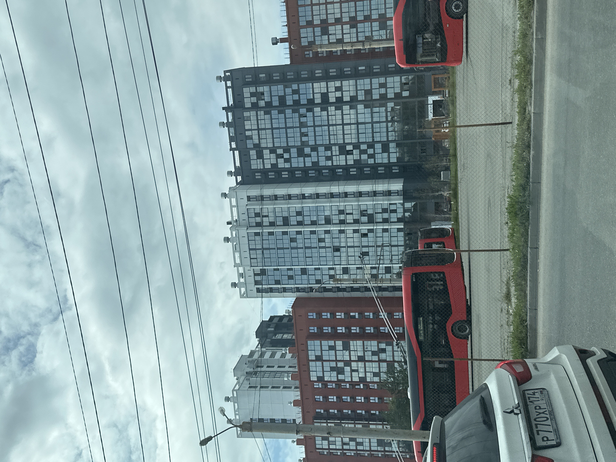An eye-level shot captures a street scene in a city. Dominating the background are several modern, multi-story apartment buildings of varying heights and colors. The buildings feature numerous windows, some reflecting the cloudy sky. In the mid-ground, two bright red buses are visible, one fully in frame and another partially. The foreground is occupied by the rear of a white car, its license plate clearly showing "P770XP 174 RUS." A chain-link fence separates the street from the area where the buses are parked, with a strip of grass and a sidewalk running along the fence. Overhead, multiple dark power lines crisscross the sky. The sky itself is filled with a mix of white and gray clouds.
