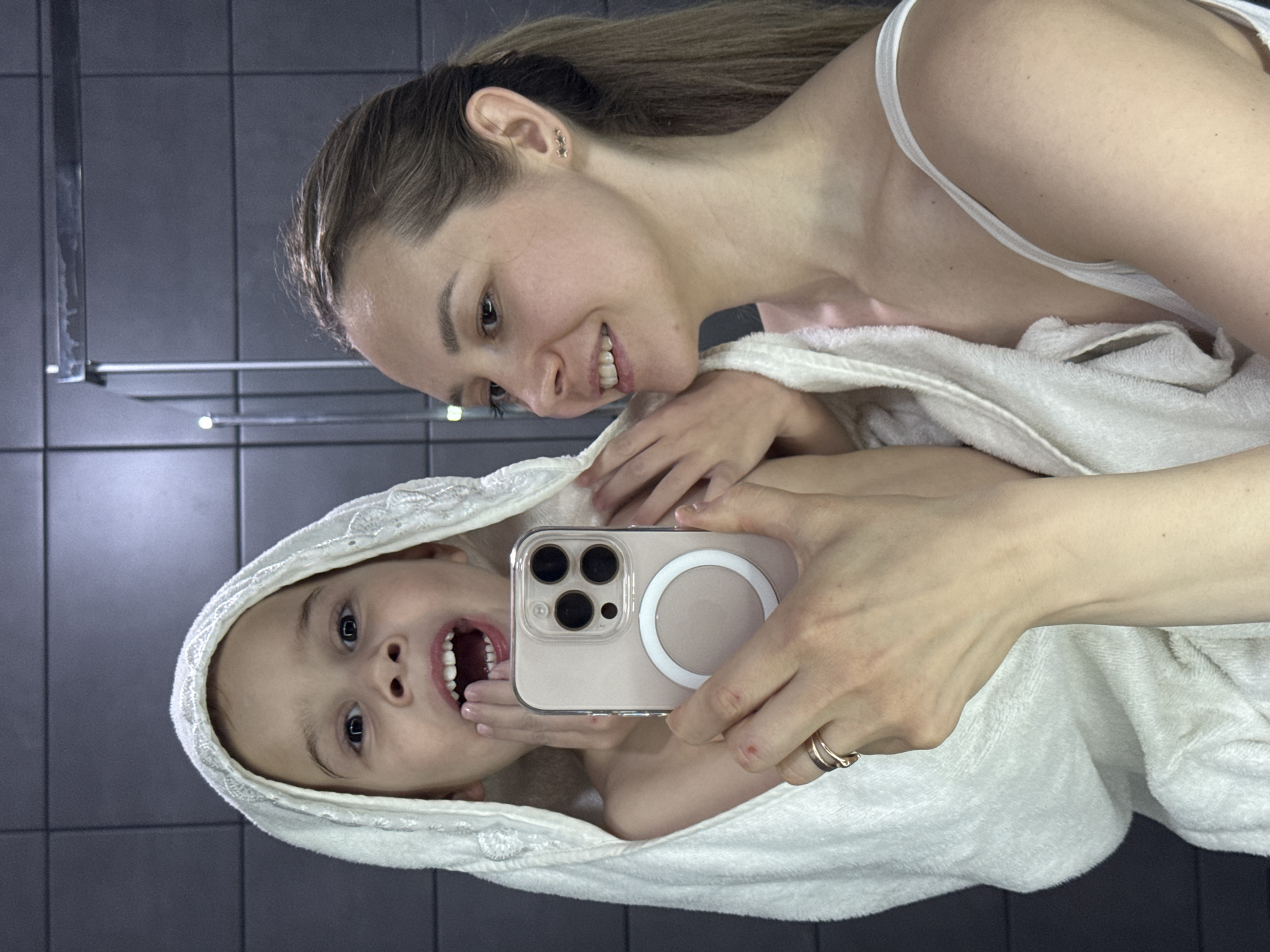 The image, rotated 90 degrees clockwise, captures a joyful and intimate moment between a woman and a child, likely in a bathroom setting in Chaikovsky, Russia. The background consists of dark grey tiled walls, with blurry reflections of metal fixtures, possibly a towel rack, and lights.
The woman, positioned above and to the right of the child (in the original image orientation), is smiling warmly and looking down affectionately. She has light brown hair pulled back and is wearing a white strap, possibly from a top, beneath a white towel wrapped around her. A small, star-shaped gold earring is visible in her right ear.
The child, wrapped in a white hooded towel, is directly facing the camera with a wide-open mouth, revealing their teeth, and bright, expressive eyes, suggesting laughter or excitement. They are holding a smartphone – an iPhone with a clear MagSafe case – indicating a selfie or a shared photograph. The woman's hand is also visible, perhaps assisting the child with holding the phone, and she wears a simple gold ring on her finger.
Both subjects appear to be in the immediate aftermath of bathing or showering, enveloped in comfortable white towels. The overall atmosphere is playful and tender, highlighting the affectionate relationship between the woman and child. No distinct text is visible in the image.