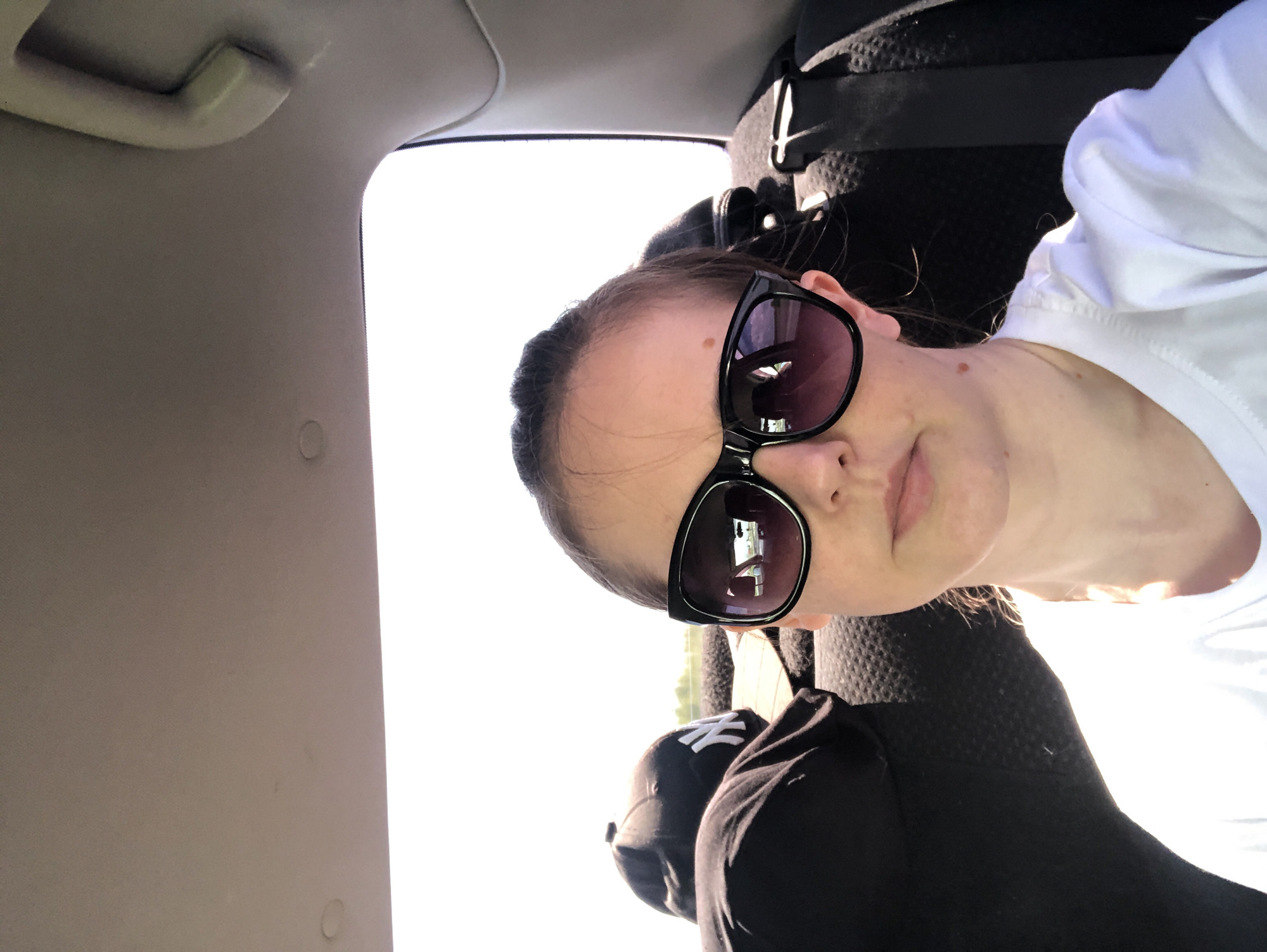 A woman with sunglasses on is taking a selfie in the back seat of a car. She is wearing a white t-shirt. To her left, a black baseball cap with a white NY logo is visible. The background is bright and appears to be outdoors.