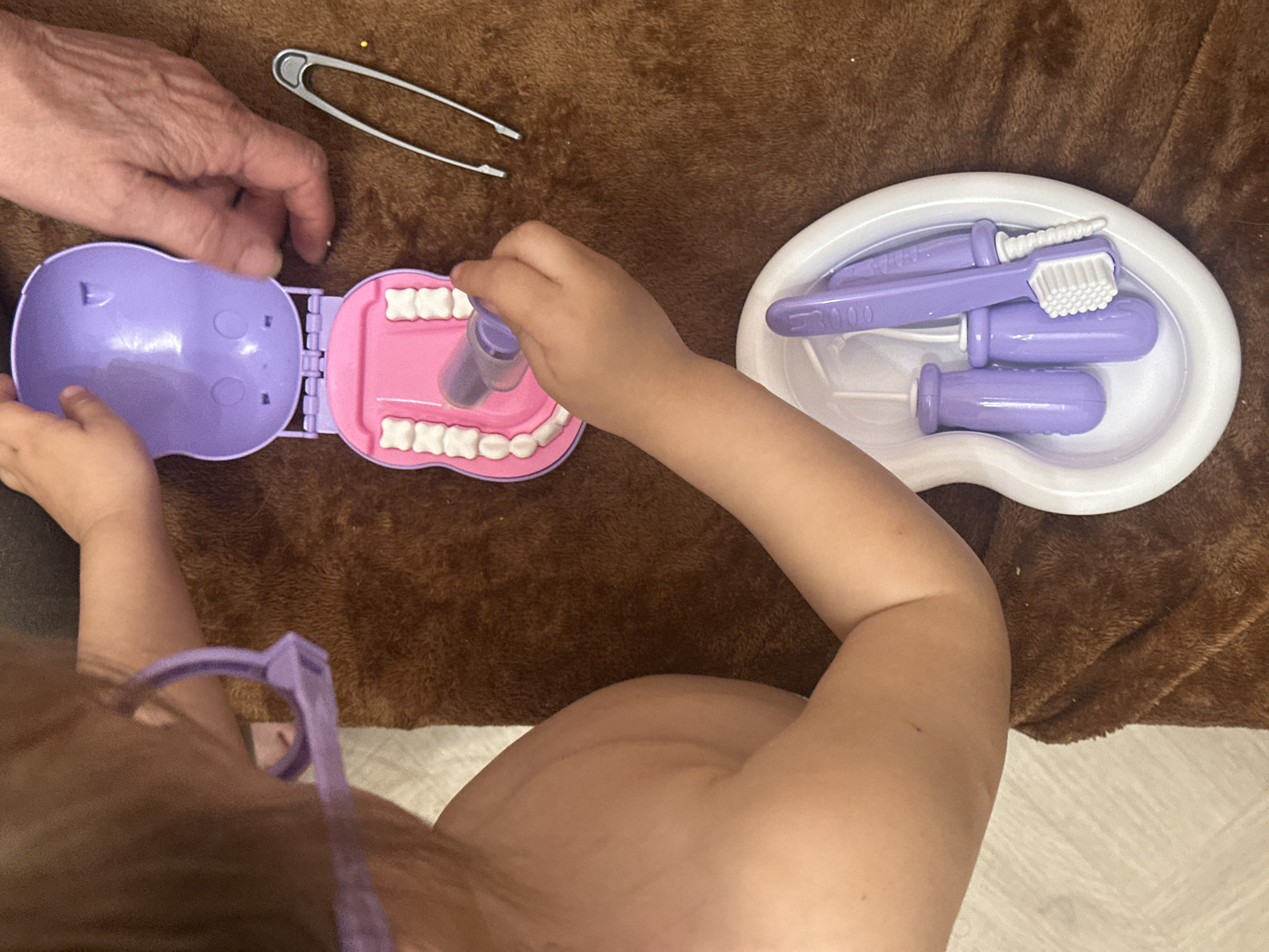 A young child, viewed from above and slightly behind, plays with a toy dentist set. The child is mostly unclothed and is wearing lavender-colored toy glasses. The dentist set, primarily in shades of lavender and white, includes a hinged plastic case resembling a mouth with plastic teeth, a white kidney-shaped tray containing several miniature dentist tools (toothbrush, probe, mirror), and a toy syringe held by the child. An adult hand assists with opening the case. A silver tweezer lies near the adult hand. The scene is set on a textured brown blanket. The location in Kharnavy, Russia, is not determinable from the image.