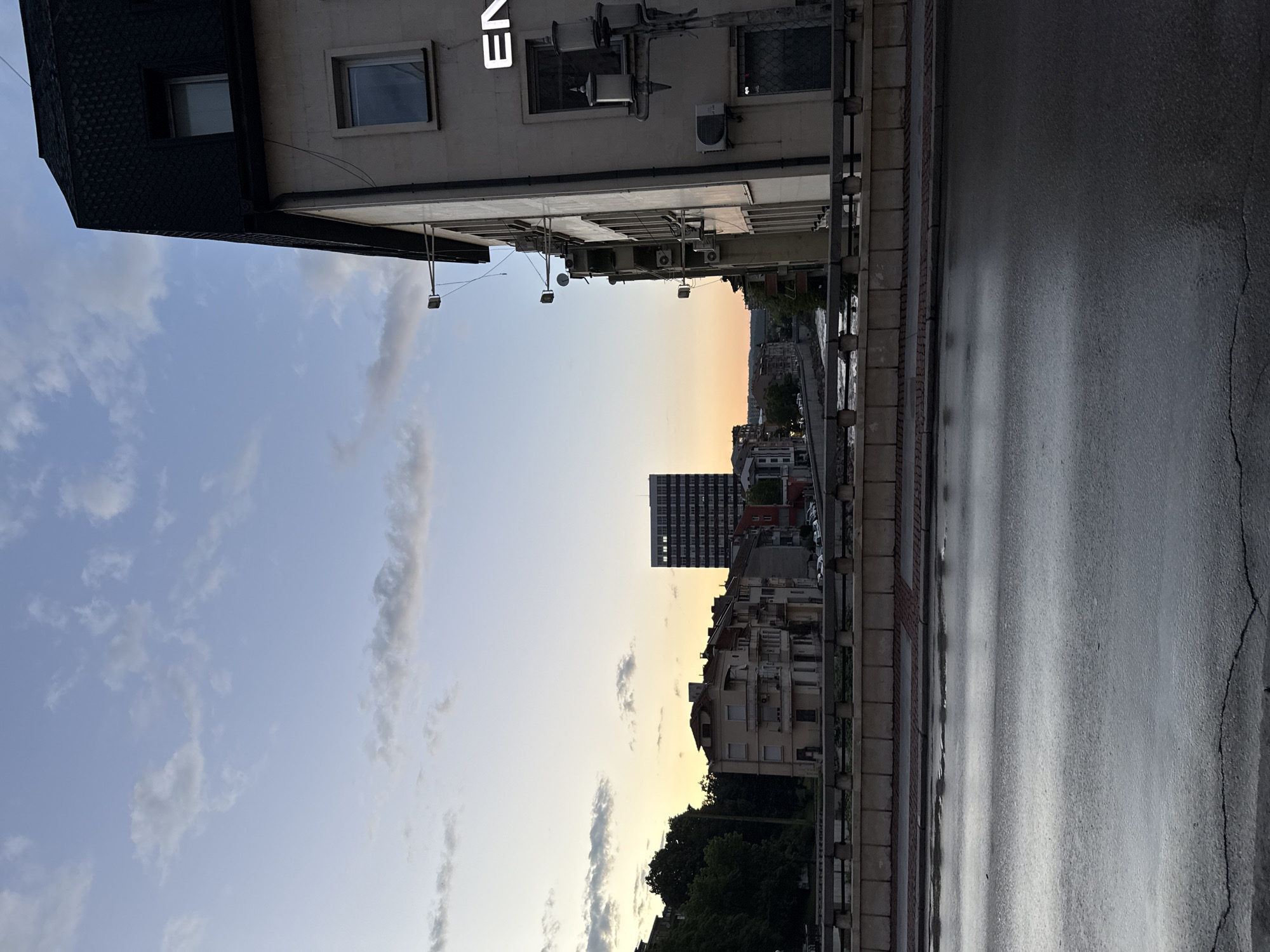The image captures an urban street scene in Gabrovo, Bulgaria, during dusk or sunset. The perspective appears to be looking down a street or alley from a slightly elevated position.
In the immediate foreground, to the left, is the corner of a building with a dark, textured facade on its upper section and a lighter, possibly cream-colored facade below. This building features several windows, multiple external air conditioning units, and visible wiring. A white, glowing sign displaying the letters "EN" is mounted on this building.
To the right in the foreground, a wet asphalt road or street surface is visible, showing reflections of the sky and ambient light. A sidewalk or curb area with what appears to be brick-like paving stones separates the building from the road. The asphalt exhibits some cracks, particularly towards the right side of the frame.
The midground and background reveal a cityscape with various buildings, including a prominent, dark, multi-story high-rise building in the distance. Clusters of trees are visible below the distant buildings, suggesting greenery within the urban environment.
The sky is a notable feature, displaying a gradual transition from a light blue with scattered white clouds on the left to warm hues of orange and yellow near the horizon on the right, characteristic of sunset or twilight. The overall atmosphere is calm and still, with no specific human activity evident. The wet road might suggest recent rain, though the sky is clear at the time of the photograph.
