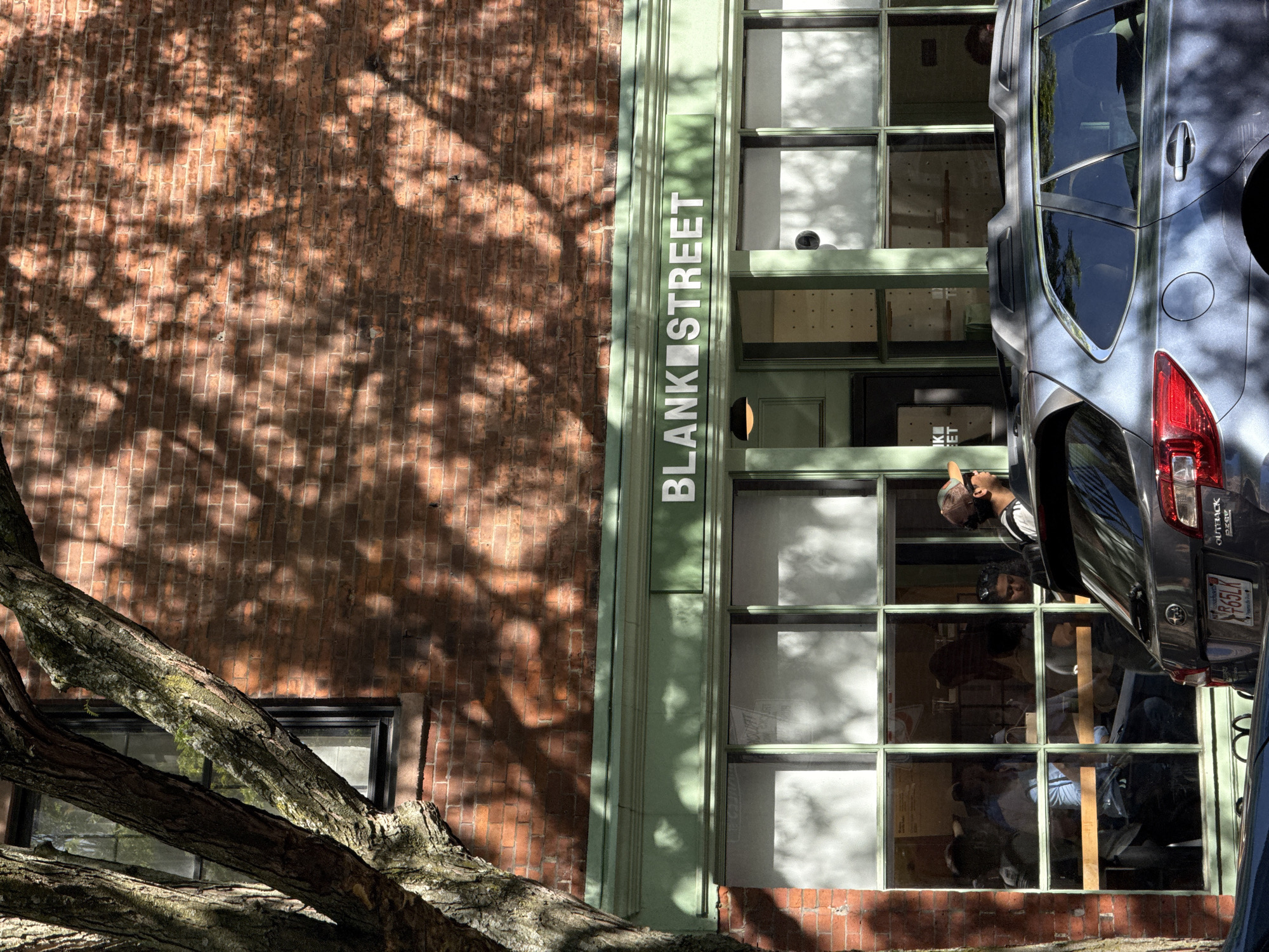The image shows the exterior of a building on "Blank Street," indicated by a sign on the building's façade. The building appears to be located on a brick street, with a red brick wall visible to the left and below. A tree with exposed branches is located in the foreground.
The building itself has a green frame around multiple large windows, some of which are reflecting light and obscuring the interior. However, in one of the windows, individuals are visible, including a person wearing a cap and looking upwards.
A gray vehicle, possibly a Subaru Outback, is parked partially obstructing the windows. The license plate is partially visible, showing "R65LK" and "Connecticut" along the bottom.
The image captures an urban environment with architectural and natural elements, and depicts people inside the building. The shadows of leaves are cast on the brick walls. There is no information to confirm that this is Boston, Massachusetts.