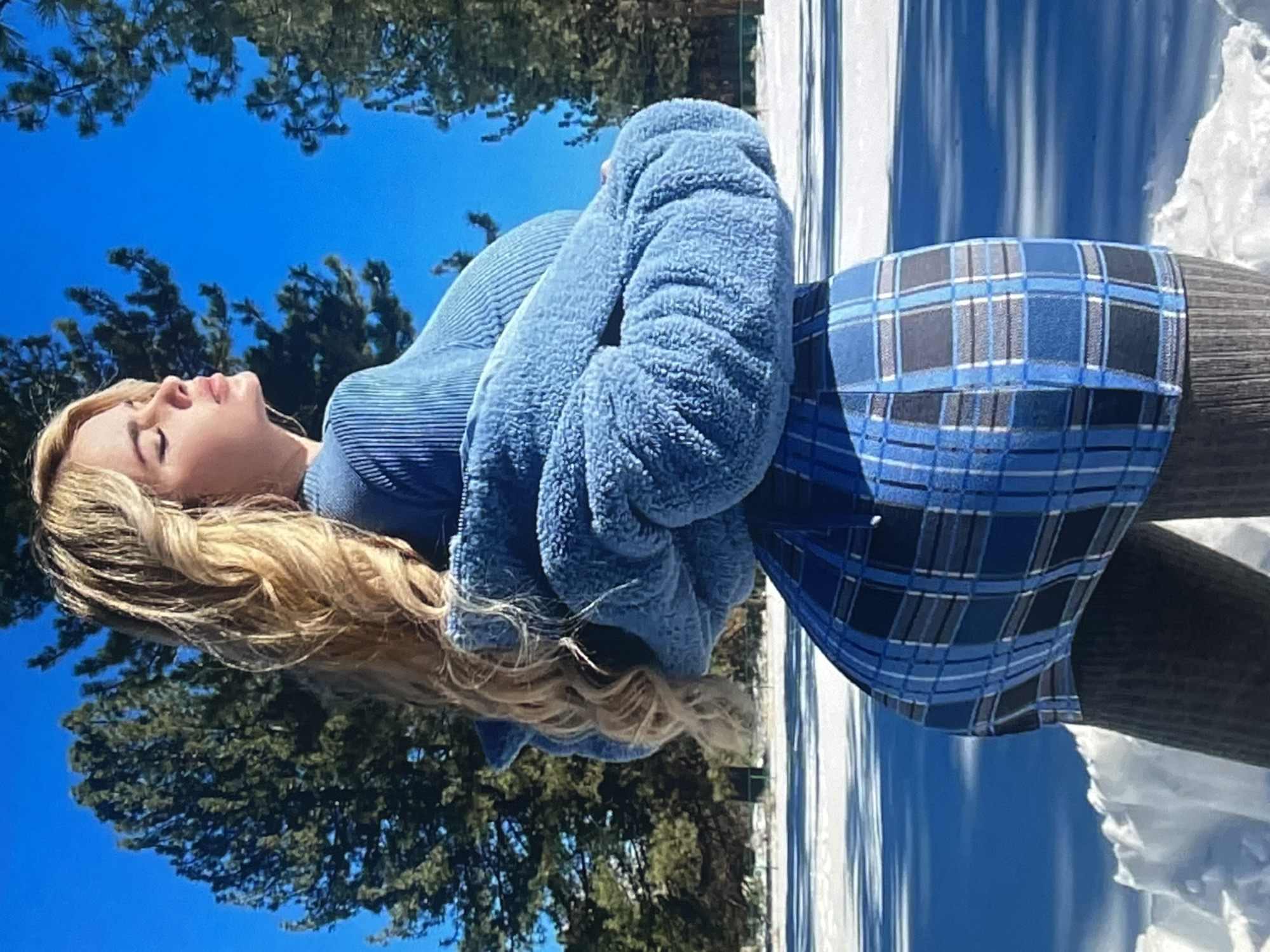 This is a vertical, eye-level, medium shot of a young woman lying down with her eyes closed and arms crossed. She has blonde, wavy hair that falls around her face and shoulders. She is wearing a blue, ribbed turtleneck sweater and a blue plaid skirt. She appears to be outdoors in a snowy environment with pine trees and a clear blue sky in the background. The snow is visible on the ground and on the pine trees. The sun is shining, creating shadows on the snow and indicating it is daytime. The woman's expression suggests she is relaxed or peaceful. There is no visible text in the image. The scene appears to be set in Vanadzor, Armenia, given the snowy conditions and pine trees that are characteristic of the region.