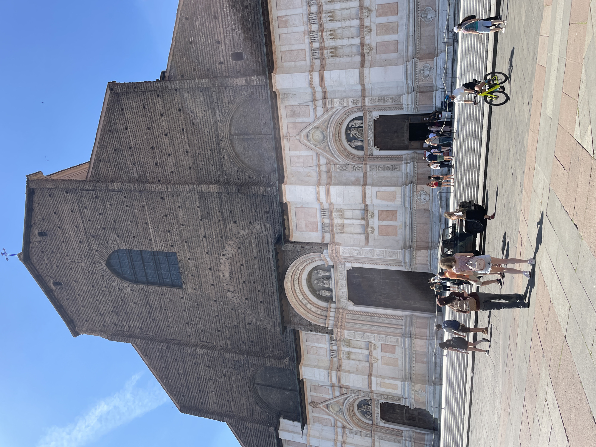 The image shows the Basilica di San Petronio in Bologna, Italy. The basilica's facade is a mix of rough brickwork and white marble. A group of people is lined up on the steps leading to the entrance. A person is riding a bicycle across the piazza in front of the church, while others are walking by. The piazza has a light brown and gray tiled surface. The sky is clear and blue.