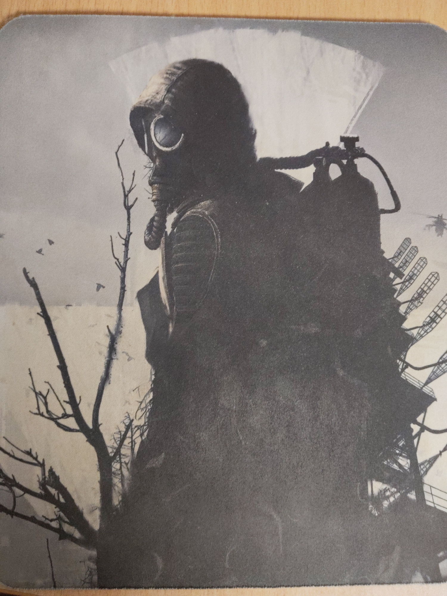 A monochromatic image depicts a figure wearing a gas mask and hooded clothing. The figure is positioned on the left of the frame, looking towards the left, with a tank attached to their back. To the left of the figure are bare tree branches. To the right of the figure is a large metallic structure which appears to be a communication tower, with a helicopter flying in the distant background. The setting is possibly an abandoned city environment. The image appears to be a photograph of a printed material, possibly a t-shirt or mousepad. There is no information to confirm the location is the city of Perm, Russia.