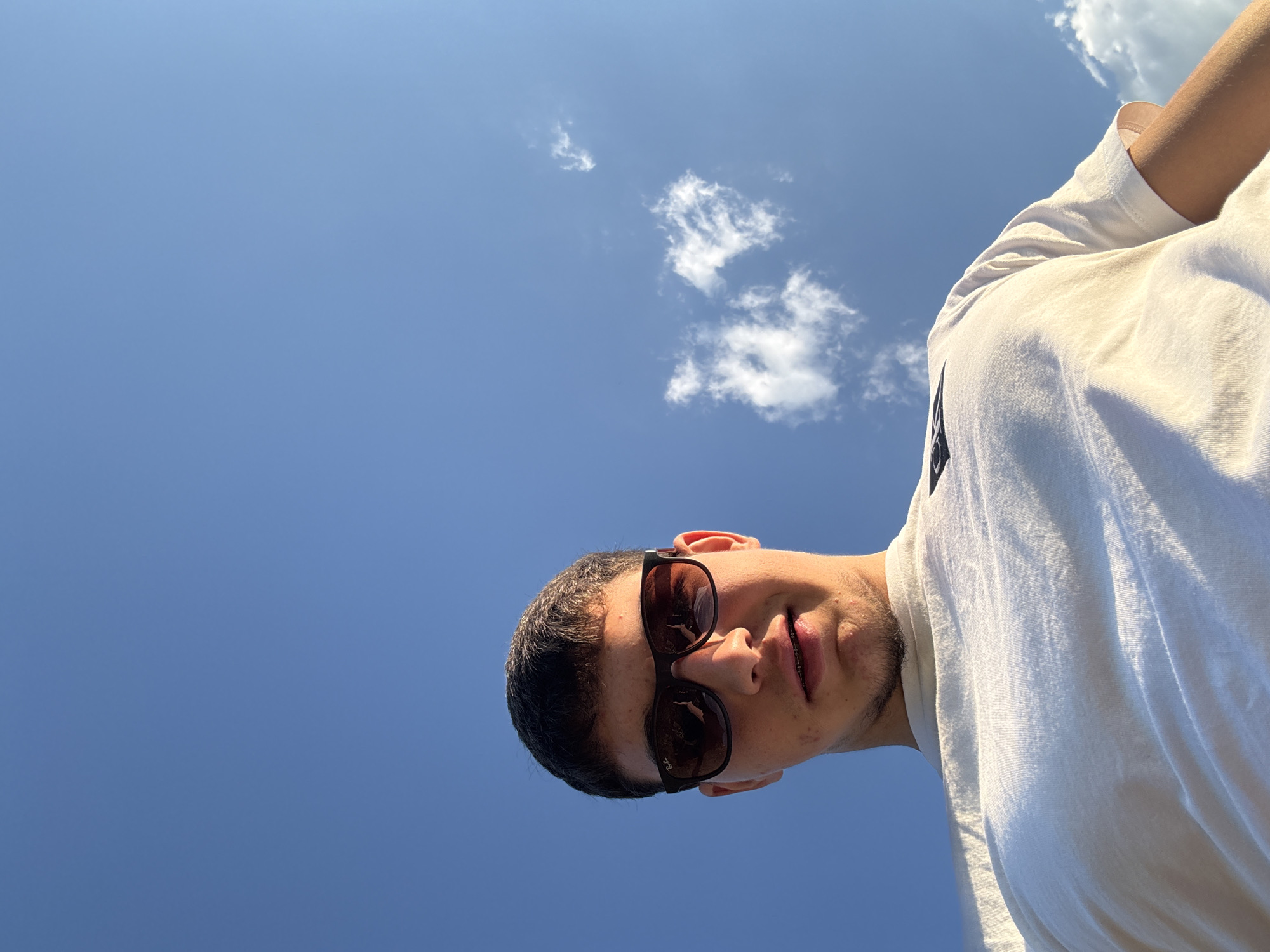 A young male with short dark hair and braces is looking up at the camera. He is wearing sunglasses and a white t-shirt with a logo on the left chest. The background is a clear blue sky with a few wispy white clouds. The image was taken from a low angle, looking up at the subject. The location is in the city of Ribartsi, Bulgaria.
