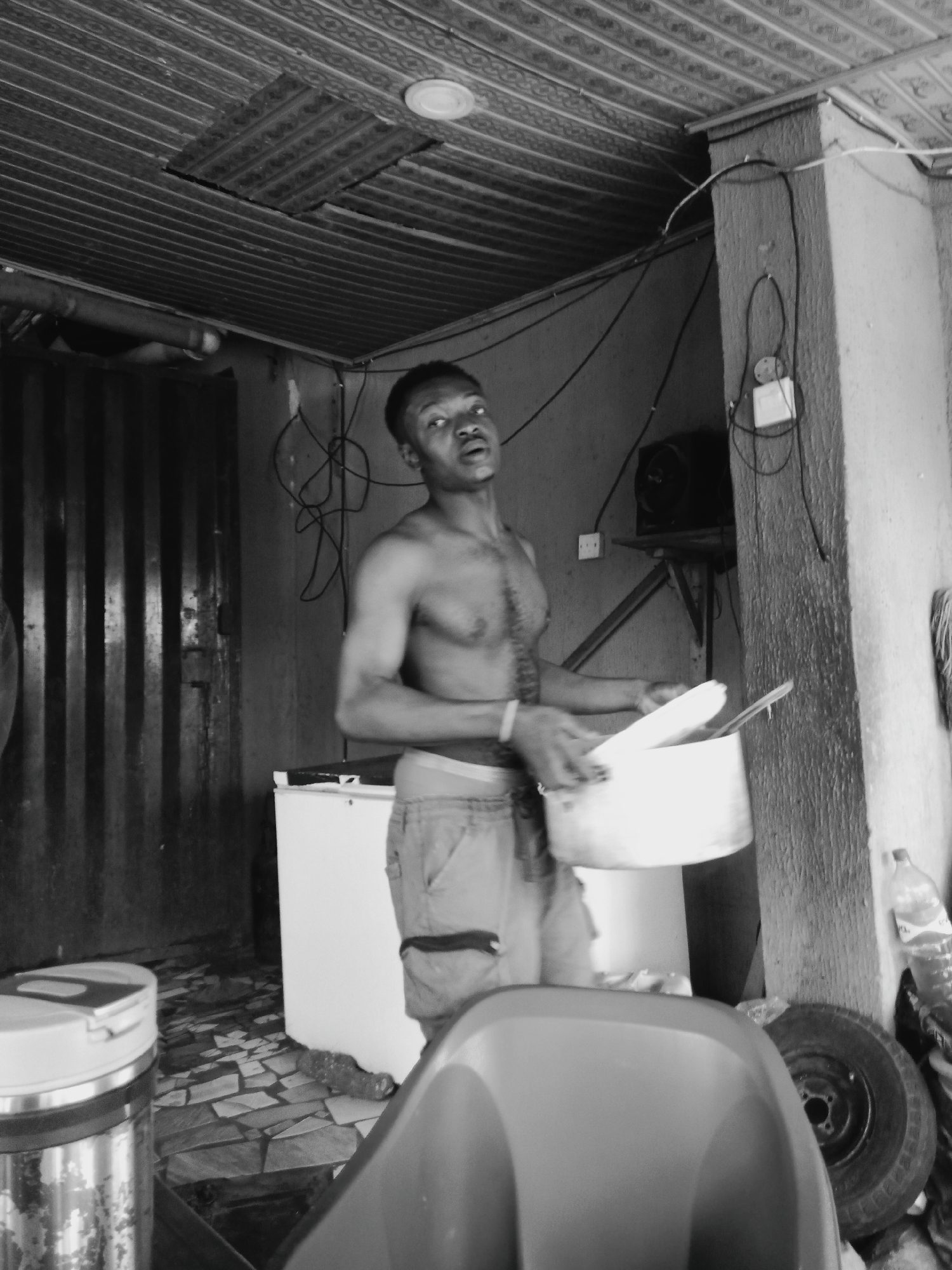 A shirtless man stands in what appears to be an outdoor kitchen in Onitsha, Nigeria. He is holding a pot and cutlery. The kitchen is makeshift, with corrugated metal roofing and unfinished walls. A refrigerator is visible behind him, and a spare tire leans against a pillar. The floor is tiled. Wires hang haphazardly from the wall and ceiling. A speaker is mounted on a shelf in the background. A chair and a metal container are in the foreground.