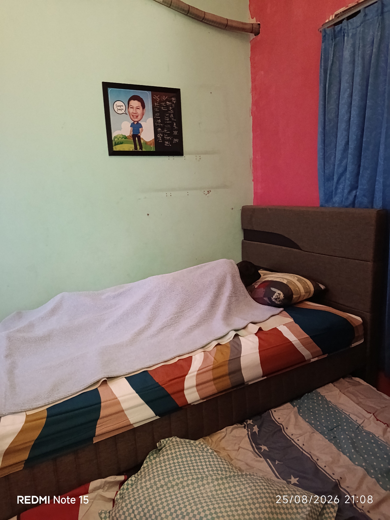 In a bedroom in Keboananom, Indonesia, a person is lying in a bed covered with a light blue blanket. The bed has a dark brown headboard and is made with a comforter featuring stripes in red, orange, brown, teal, and white. A pillow with a striped pattern in shades of brown and yellow is visible. Another bed or mat on the floor is covered with a comforter adorned with blue stars on a white and blue striped background, and a green and white checkered pillow. On the light green wall above the bed, a framed picture is hung. The frame contains a caricature of a man with a speech bubble that reads "Sampai jumpa," meaning "See you later." To the right of the caricature, there is a section with what appears to be text or symbols. The walls of the room are painted light green on the left and a bright pink on the right. A blue curtain hangs on the right side of the frame, partially obscuring a window. A copper pipe runs along the top of the wall. The timestamp "25/08/2026 21:08" and "REDMI Note 15" are visible at the bottom of the image.