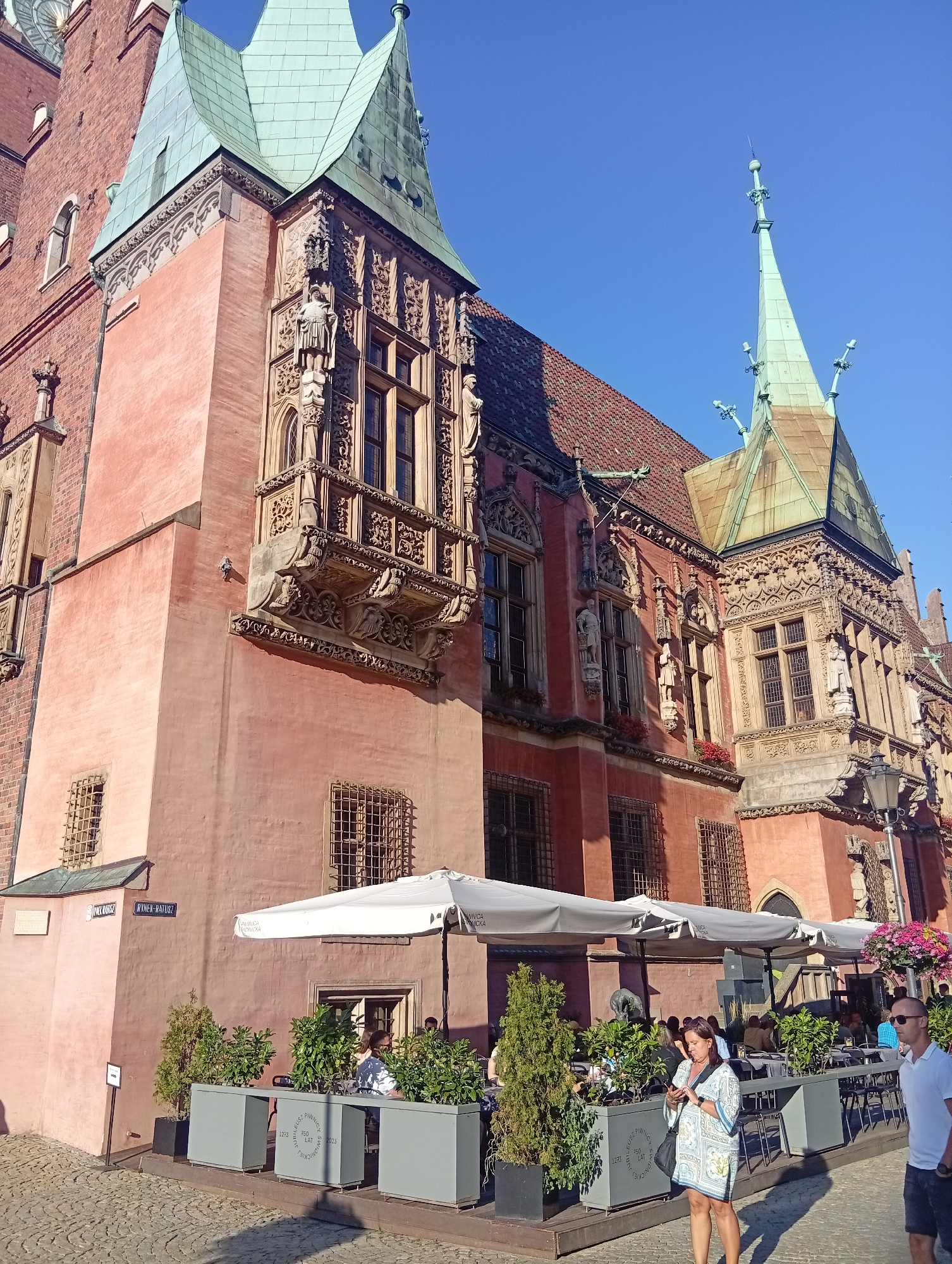The image shows the Old Town Hall in Wrocław, Poland, a historic building with a red-orange facade and ornate architectural details. The building features multiple towers with green tiled roofs, decorative carvings around windows and balconies, and large arched windows. In the foreground, there is an outdoor seating area for a cafe with white umbrellas providing shade over tables and chairs. Several people are visible: a woman in a light-colored patterned dress is looking at her phone, and a man in a white polo shirt and shorts is walking past. Lush green plants in large grey planters line the walkway in front of the cafe. The ground is paved with cobblestones. The sky is clear and blue, indicating a sunny day. Signs with Polish text are visible on the building's exterior.