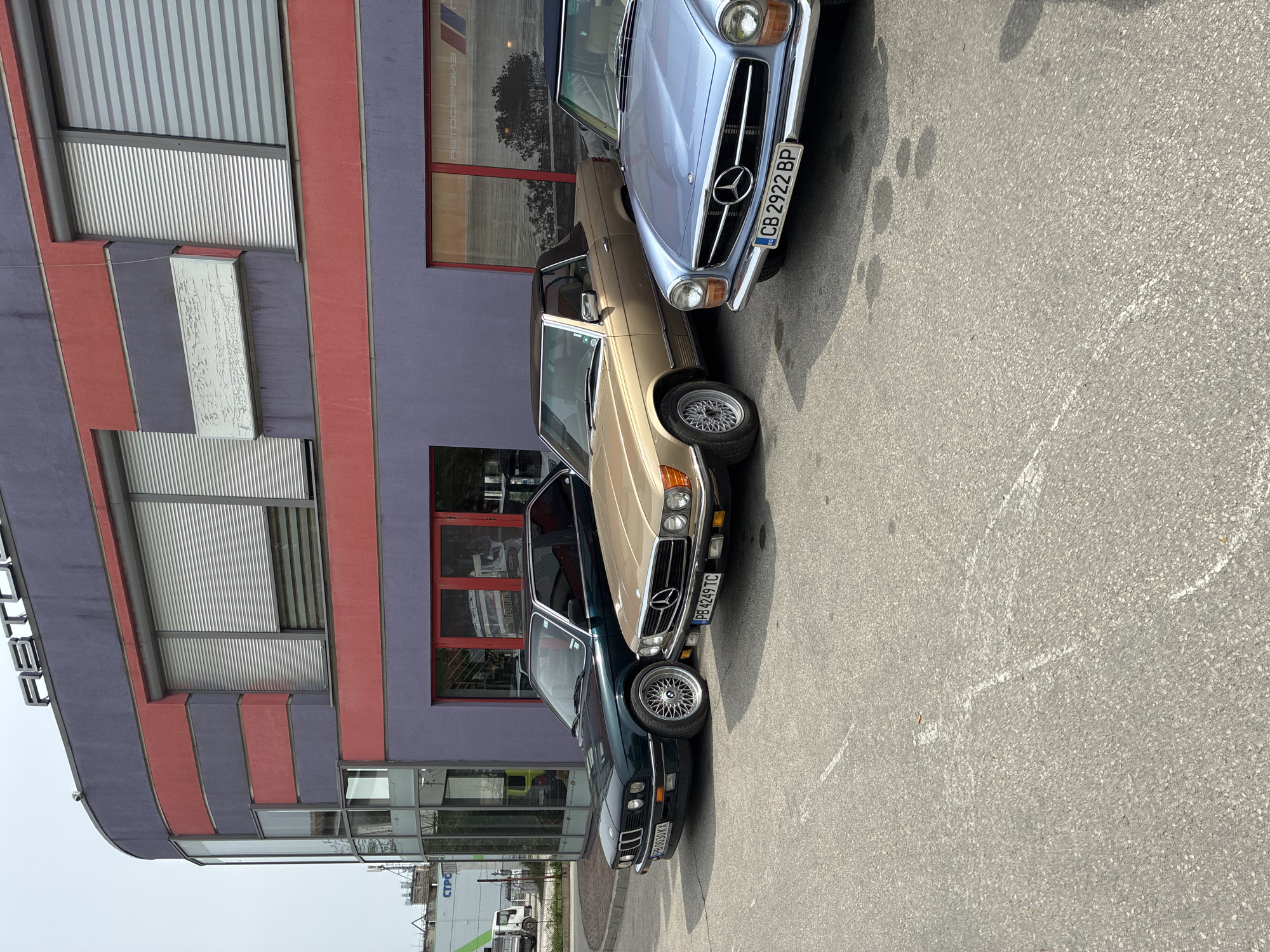 The image captures an outdoor scene featuring three classic cars parked in front of a modern building, likely in Plovdiv, Bulgaria. The time of day appears to be bright and sunny, indicated by clear lighting and shadows on the asphalt ground.
The primary subjects are three vintage cars, neatly aligned. From top to bottom in the original image (or left to right if rotated correctly):
1. A light blue Mercedes-Benz R107 SL, identifiable by its distinct grille with the Mercedes star and round headlights. It displays a Bulgarian license plate: "CB 2922 BP".
2. A gold or tan Mercedes-Benz R107 SL, possibly with a hardtop, featuring multi-spoke BBS-style wheels. Its Bulgarian license plate is partially visible as "PB 4249 TC".
3. A dark green or blue BMW E30 3-series convertible, also equipped with multi-spoke BBS-style wheels. Its Bulgarian license plate reads "PB 0030 XY".
The cars are parked on an asphalt surface, which shows some faded white markings and dark stains. They are positioned facing a multi-story building characterized by a blend of purple and red paneling, giving it a contemporary, almost industrial, aesthetic. The building features large windows, some with horizontal blinds. Reflections in the windows reveal trees and parts of an interior space. One window clearly reflects Cyrillic text, appearing to spell "АВТОСЕРВИЗ" (AVTOSERVIZ), which translates to "Car Service," suggesting the location might be an automotive service center or dealership specializing in classic cars. Another faint Cyrillic text, possibly "СТРО..." (STRO...), can be seen on a distant sign or building through a lower window.
No people are visible in the image. The arrangement of the well-maintained classic cars outside what appears to be a car service suggests either a display, a gathering of enthusiasts, or cars undergoing maintenance.