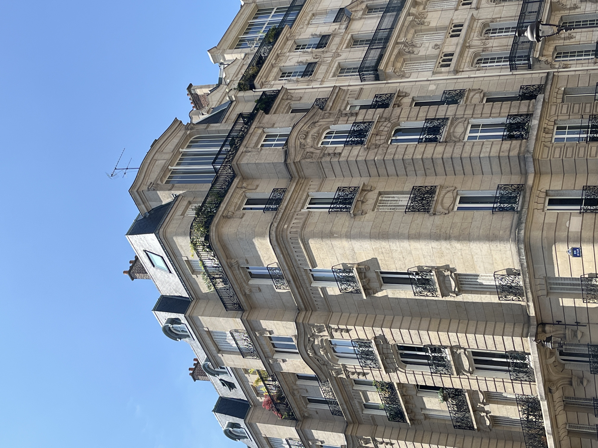 This is a low-angle, exterior, daytime, medium shot of Haussmannian apartment buildings in Paris, France, under a clear blue sky. The buildings are constructed of light-colored stone, with mansard roofs covered in dark slate. The facades feature numerous windows, many with ornate black wrought-iron balconies. Some balconies are adorned with potted plants and greenery, adding touches of color to the stone structures.
The building on the left has a rounded corner and displays intricate stone carvings above some of the windows. The building to its right is slightly set back and also exhibits similar architectural elements. Antennae are visible on the rooftops of both buildings. The overall impression is of classic Parisian architecture, with a focus on the verticality of the buildings and the decorative details of the balconies and stonework. No people or activity are depicted in the image.